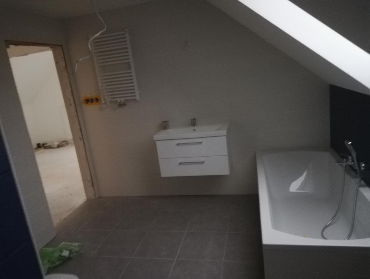 Łazienka w trakcie remontu w Nasielsku, widoczna wanna, umywalka z szafką, grzejnik drabinkowy i szare płytki podłogowe. Sufit ze skosem.