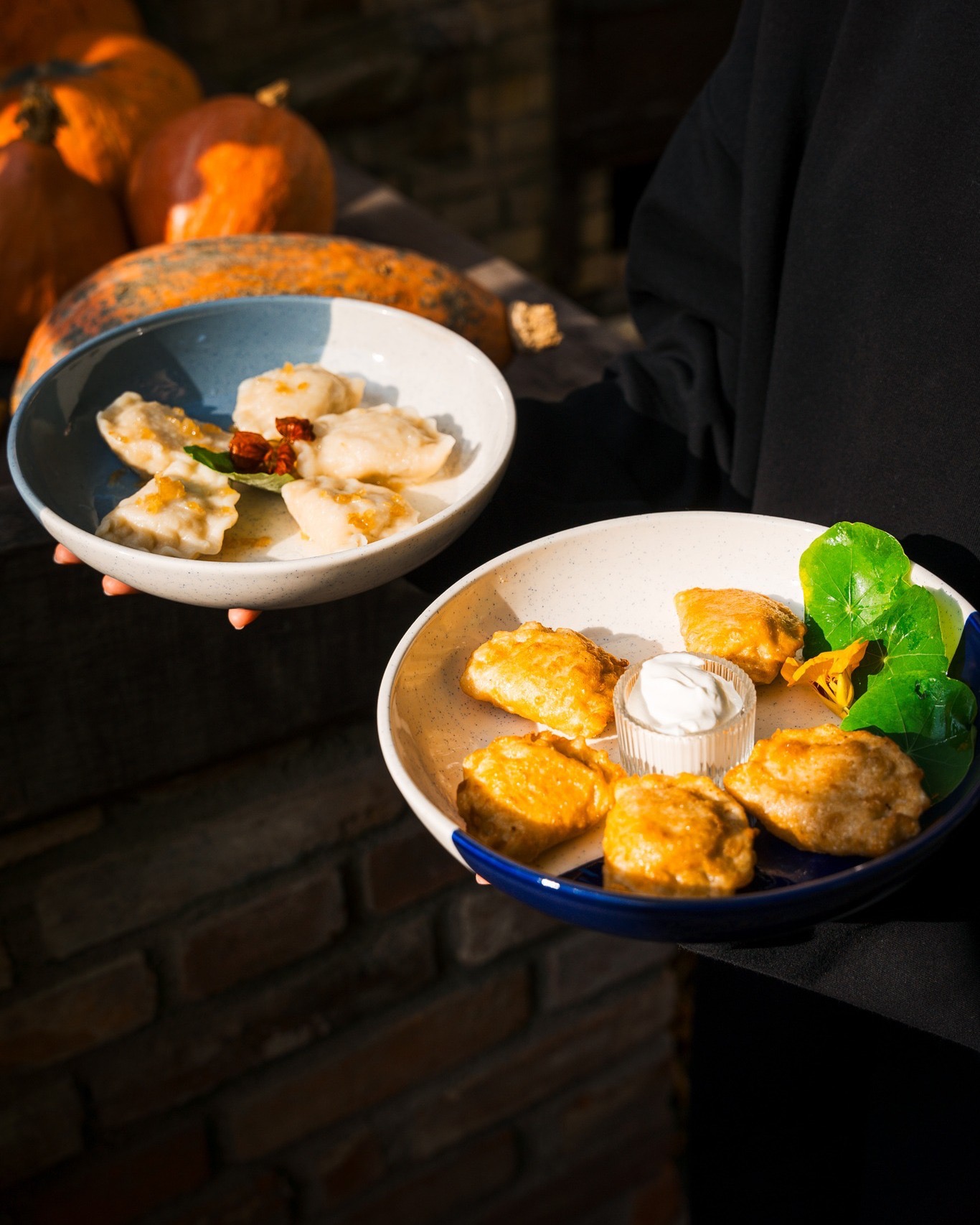 Dwie miski z pierogami: gotowane i smażone, podane z sosem i dekoracją z nasturcji. W tle dynie i ceglana ściana, ciepłe światło.