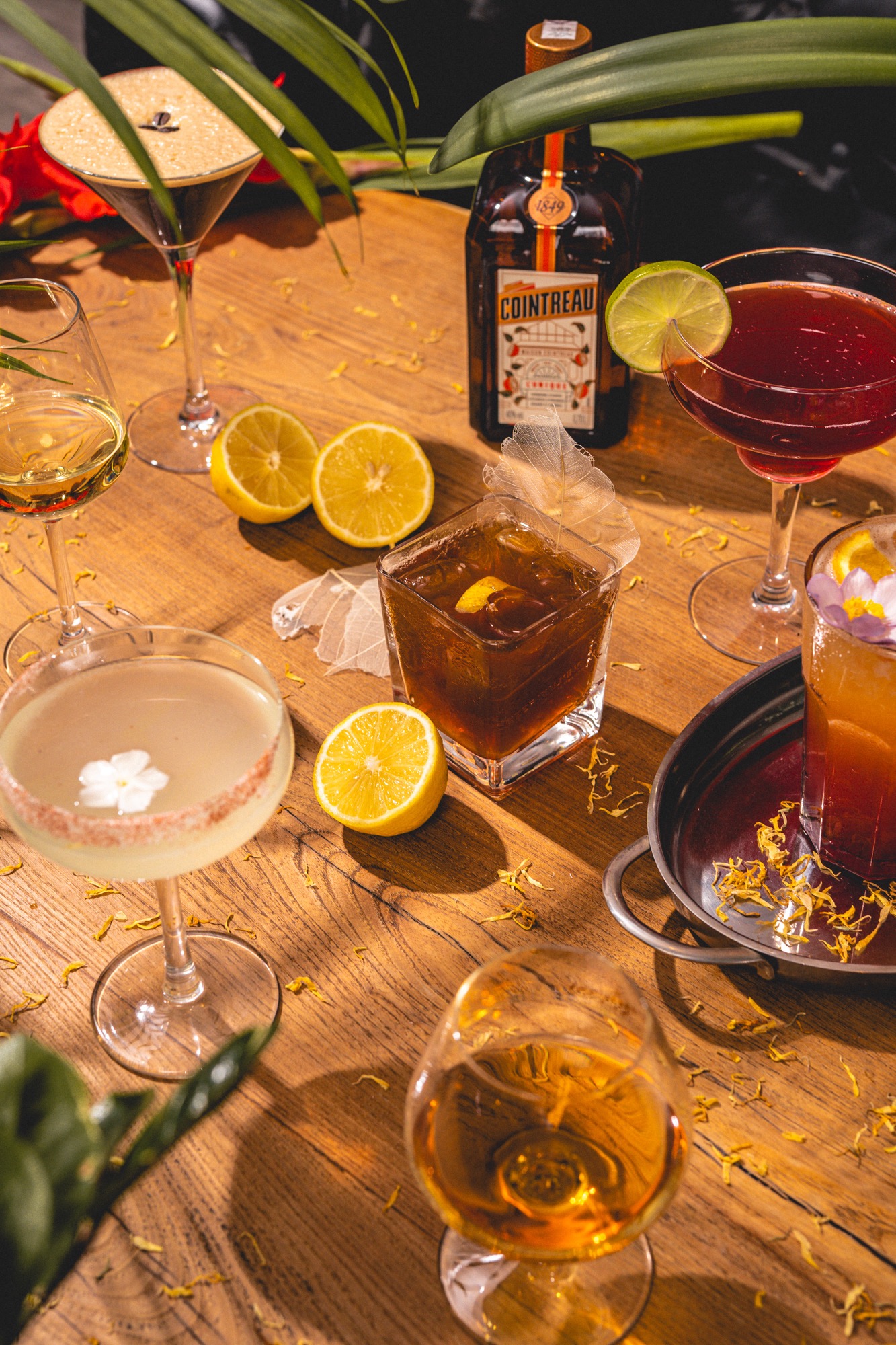 Artystyczne ujęcie różnorodnych koktajli na drewnianym stole, z dekoracjami z kwiatów i owoców, butelka Cointreau w tle. Idealne na event lub catering.