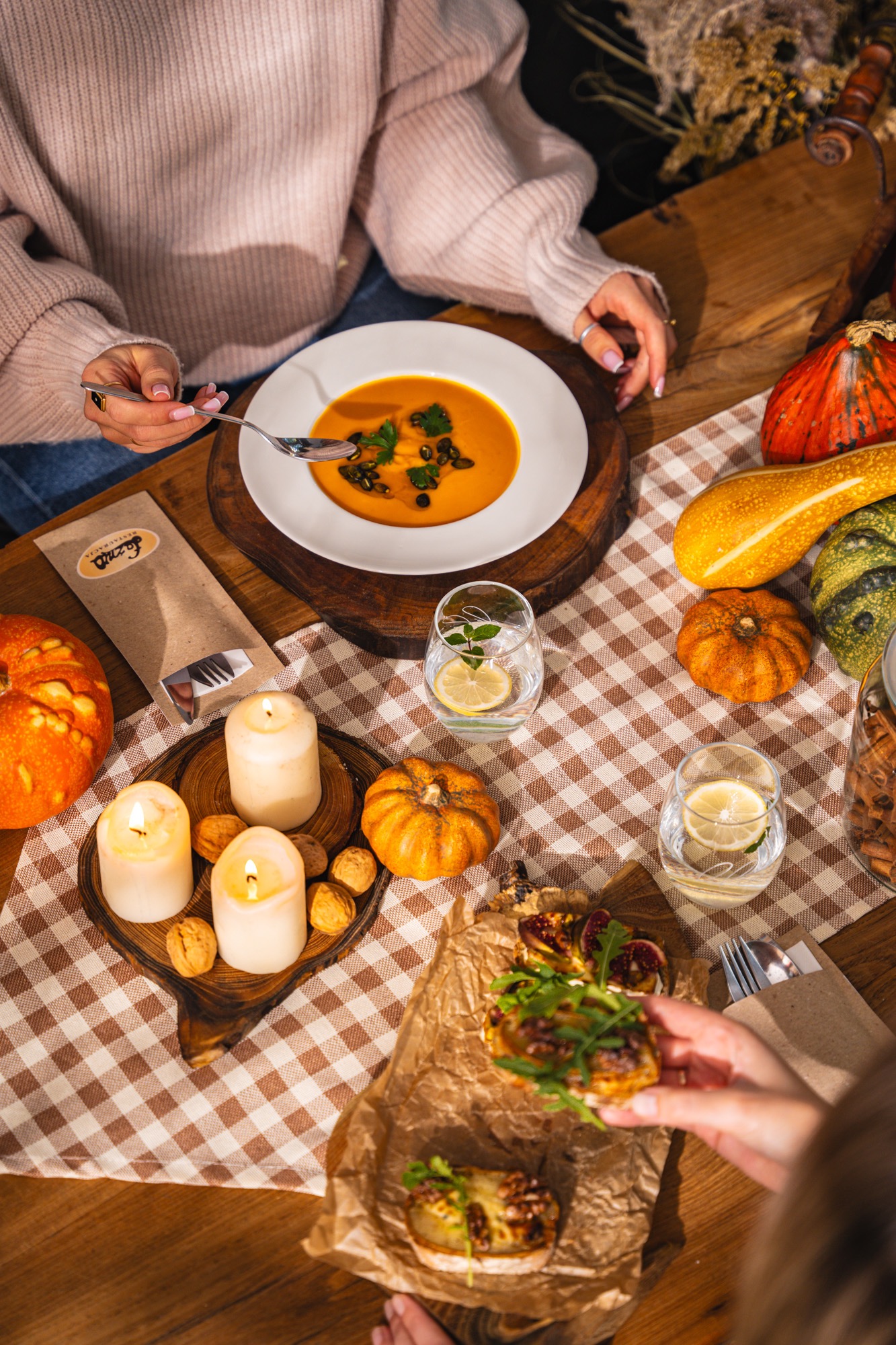 Jesienna aranżacja stołu z zupą dyniową, świecami, orzechami i kanapkami na kraftowym papierze. Dekoracje z dyń i napoje z cytryną. Ciepłe, naturalne światło.