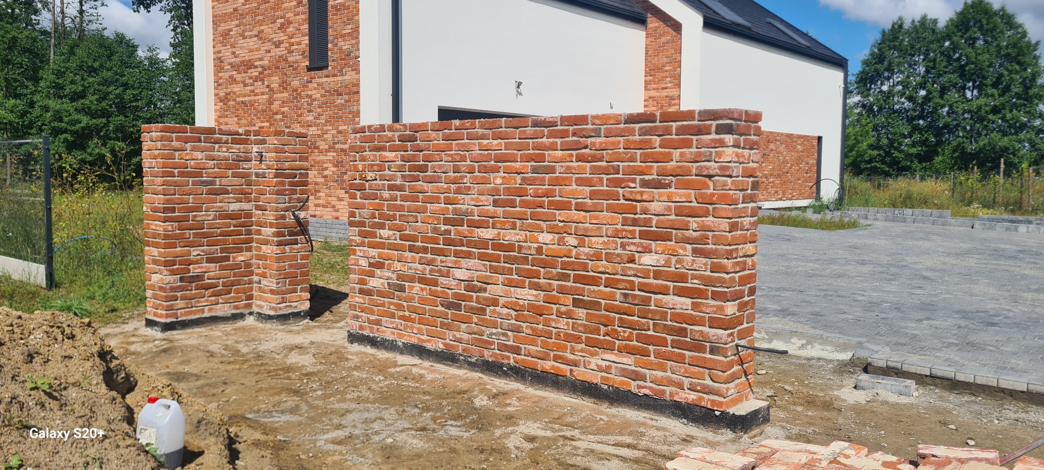 Wznoszona z czerwonej cegły ściana ogrodzeniowa na etapie budowy, widoczne fundamenty i niezabezpieczony teren wokół, w tle fragment domu z białą elewacją i ceglanym wykończeniem.