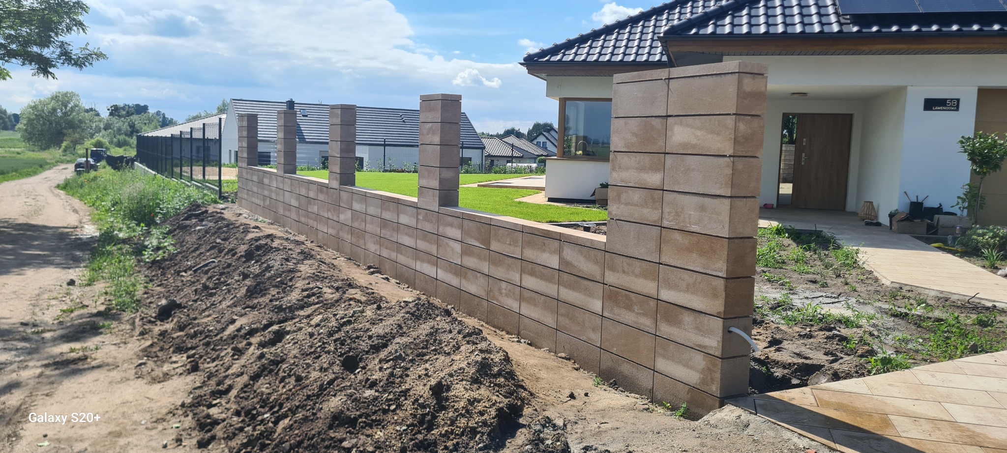 Nowoczesny, modułowy ogrodzenie z beżowych bloczków betonowych w trakcie budowy, widoczny nasyp ziemi i fragment domu z panelami słonecznymi w tle.