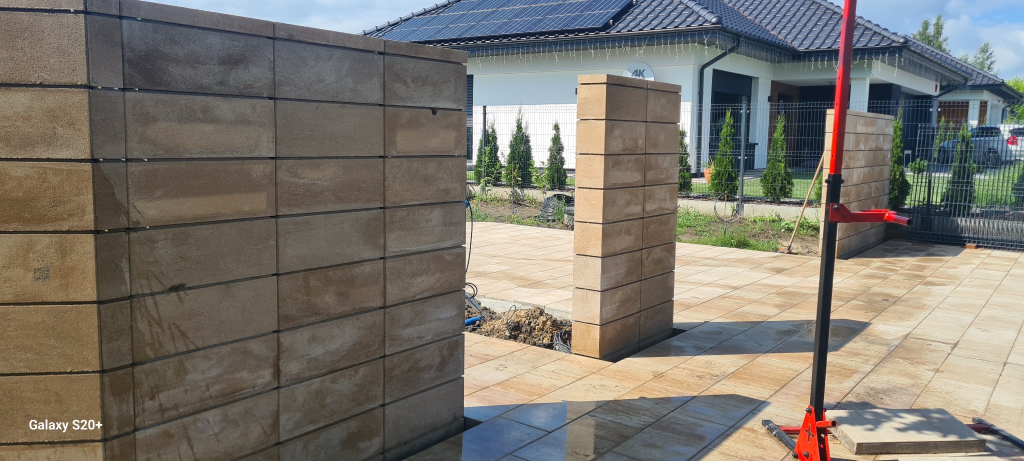 Wznoszenie ogrodzenia z betonowych bloczków modułowych; widoczne słupki i fragment muru, podłoże z kostki brukowej, w tle dom z panelami słonecznymi na dachu.