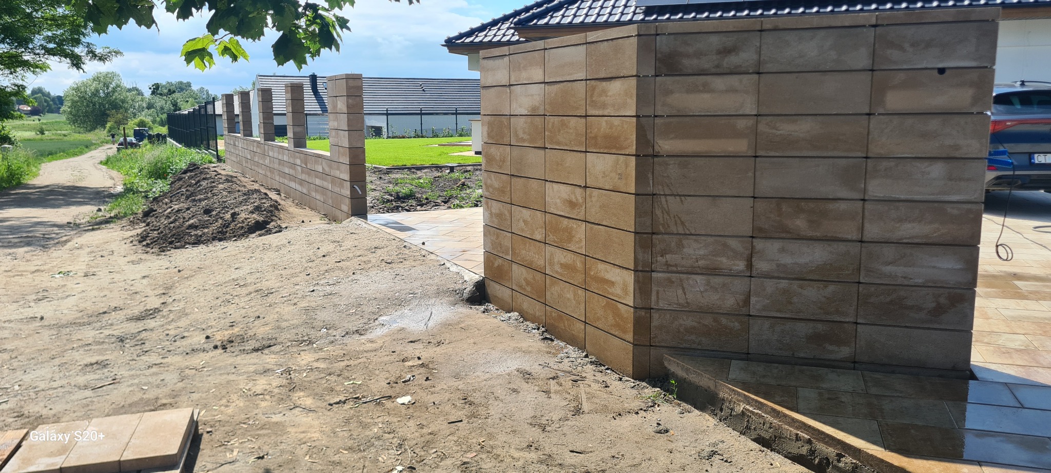 Nowoczesne ogrodzenie z bloczków betonowych w kolorze piaskowym, częściowo ukończone, z widocznym podjazdem z kostki i przygotowanym terenem pod dalsze prace.