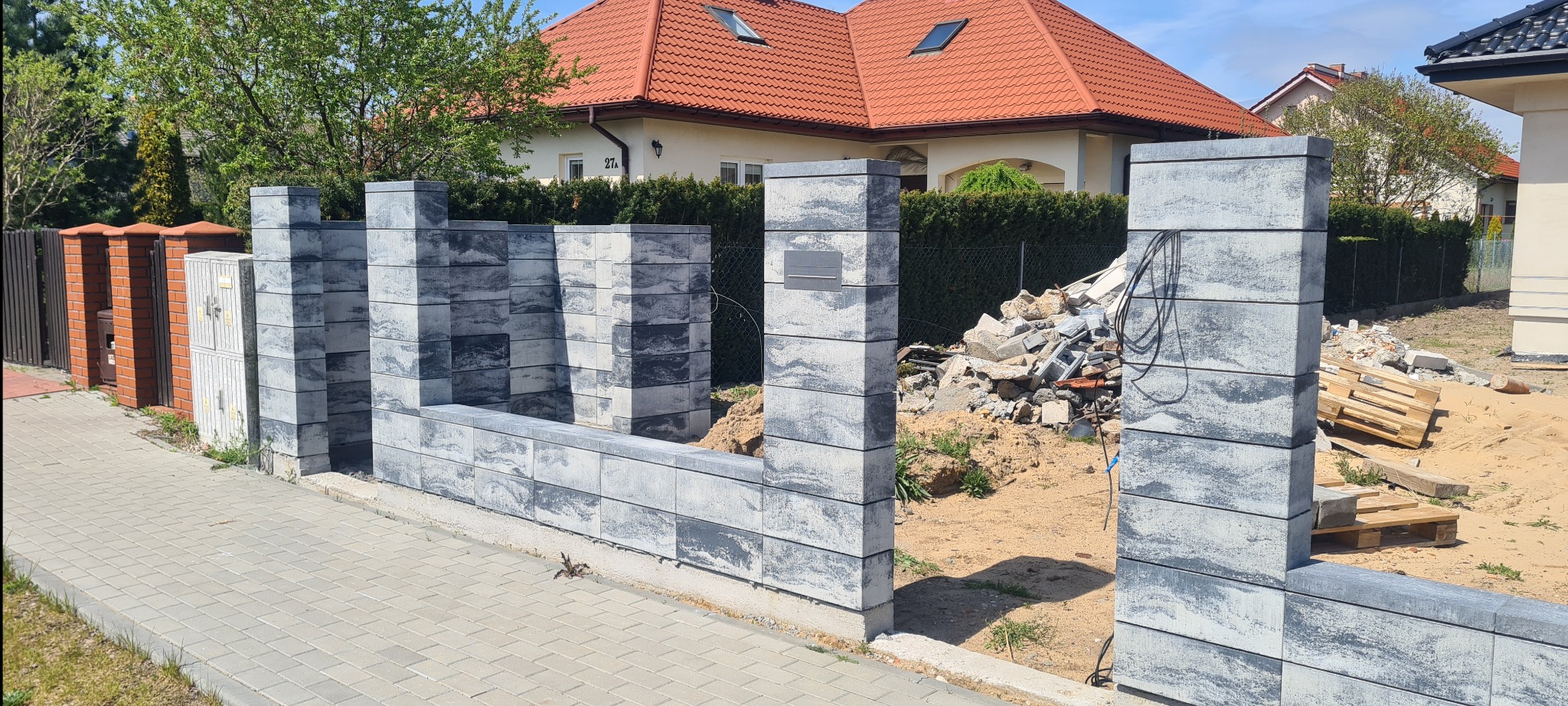 Wznoszenie ogrodzenia z szarej kostki betonowej w trakcie budowy, widoczne gruz i palety na posesji, dom jednorodzinny w tle.