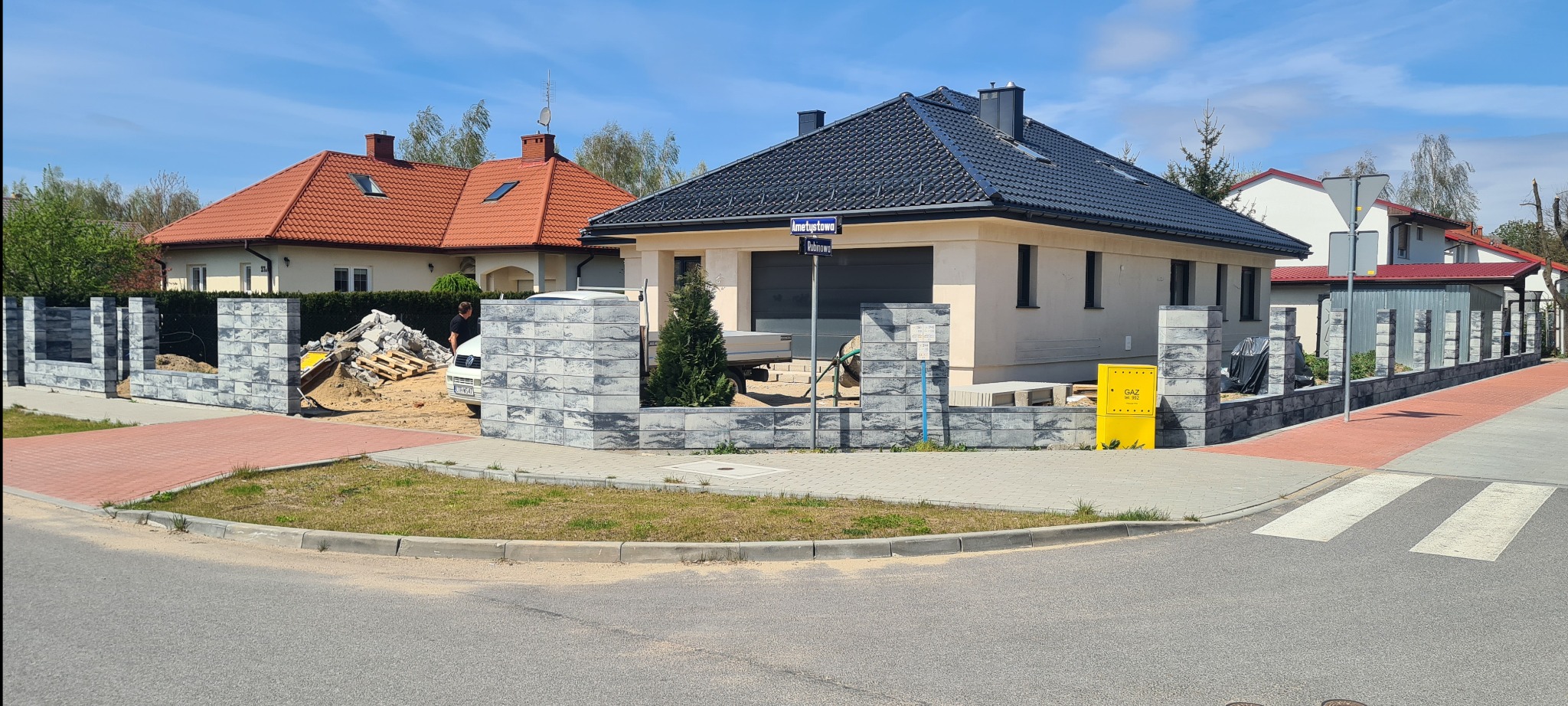 Nowoczesny dom jednorodzinny z szarą dachówką i elewacją w trakcie montażu ogrodzenia z betonowych bloczków o fakturze kamienia, widoczny żółty skrzynka gazowa i ulica z kostki brukowej.
