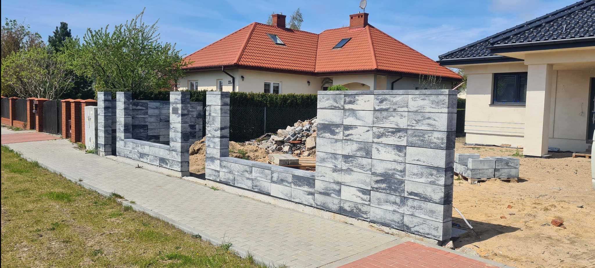 Montaż nowoczesnego ogrodzenia modułowego z szarego betonu architektonicznego w Głogowie, widoczne fragmenty domu z czerwoną dachówką i kostka brukowa.