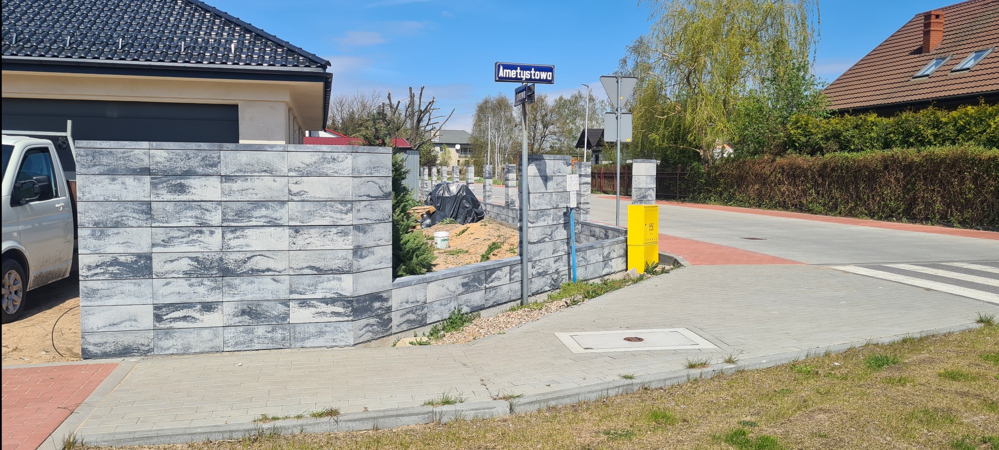 Nowoczesne ogrodzenie z szarej kostki łupanej, częściowo ukończone, z widocznym przygotowanym terenem pod dalszą budowę, obok skrzynka gazowa i znak drogowy.