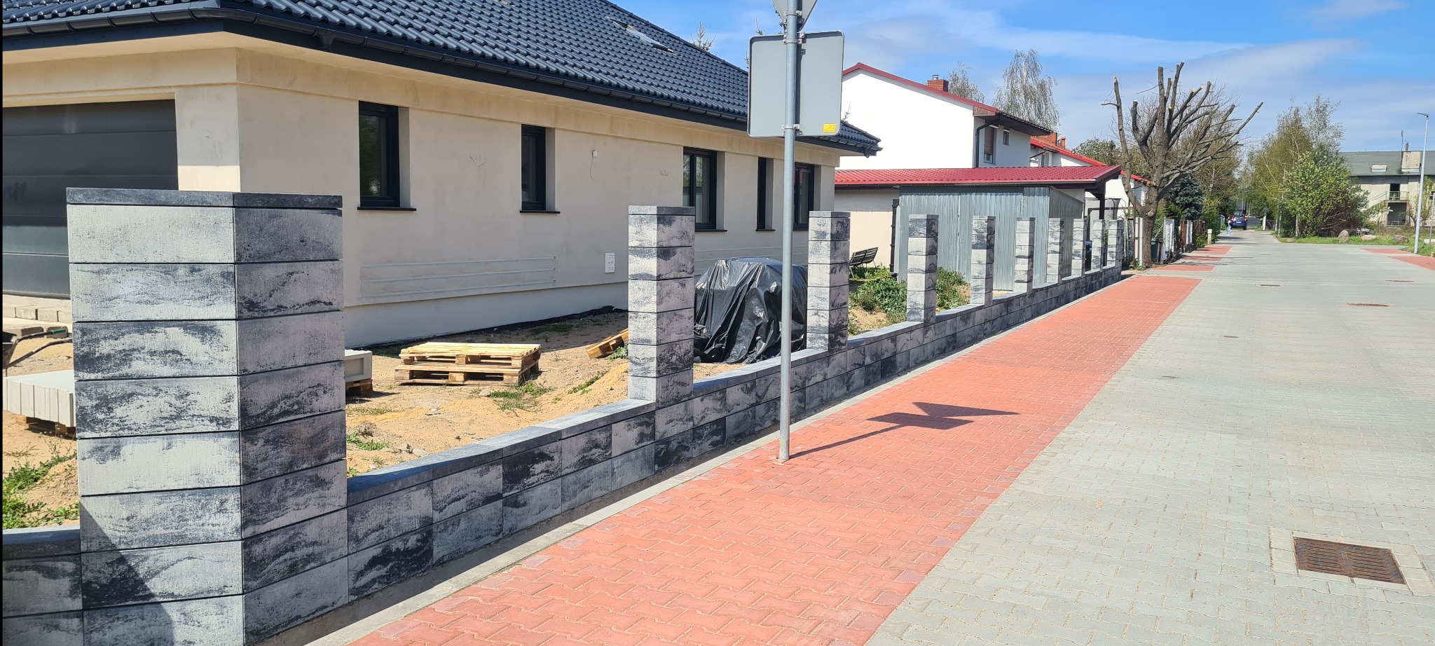 Nowoczesne ogrodzenie z szarej kostki łupanej, częściowo ukończone, z paletą i czarną folią zabezpieczającą, na tle jasnego domu z ciemną dachówką i chodnika z czerwonej kostki.