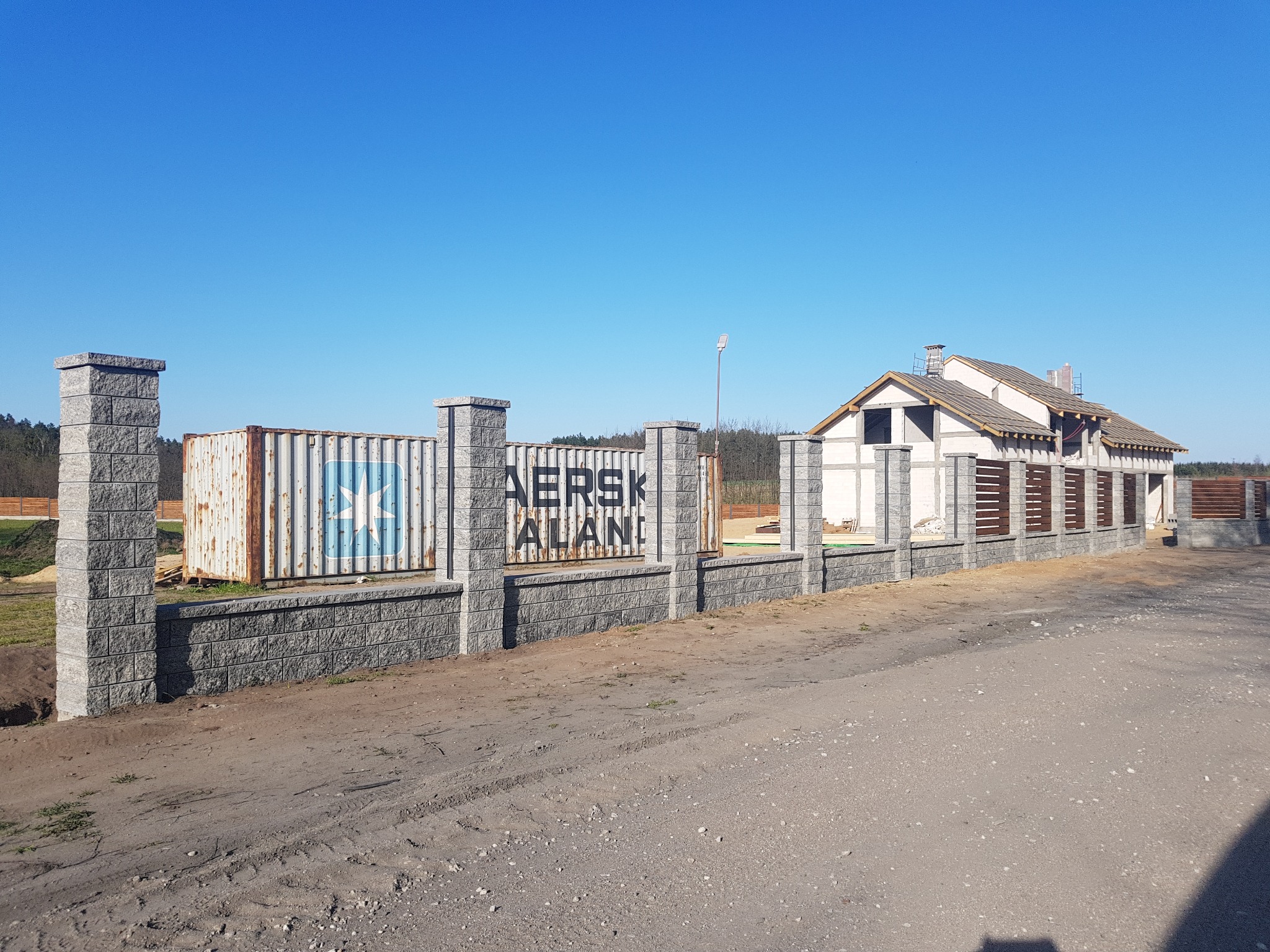 Nowoczesne ogrodzenie z szarej kostki z filarami, częściowo zasłaniające kontener transportowy i budynek w stanie surowym otwartym pod błękitnym niebem.