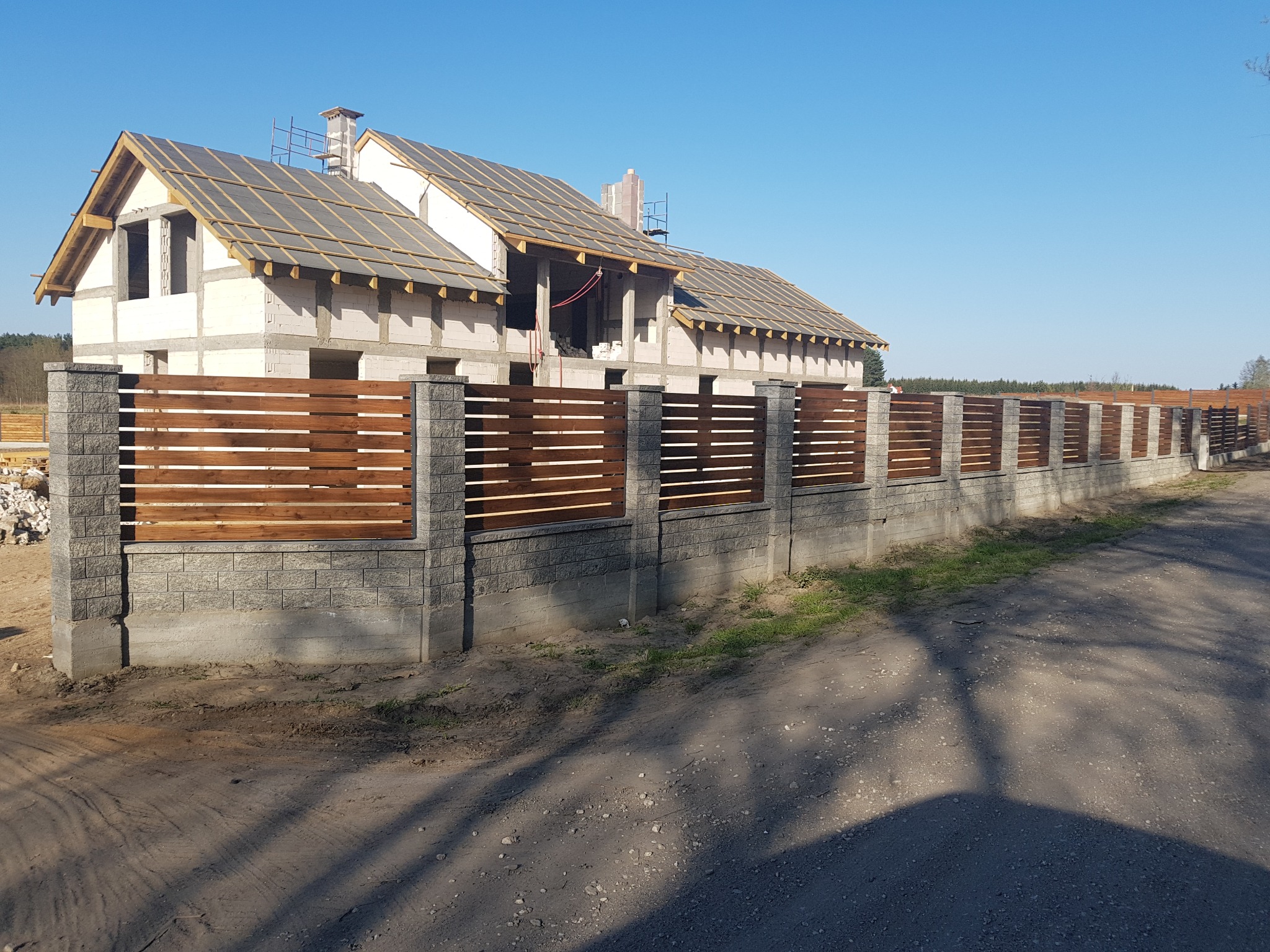 Wznoszony dom jednorodzinny z szarej cegły z zamontowanym, poziomym, drewnianym ogrodzeniem w kolorze brązowym. Widoczne rusztowania na dachu.