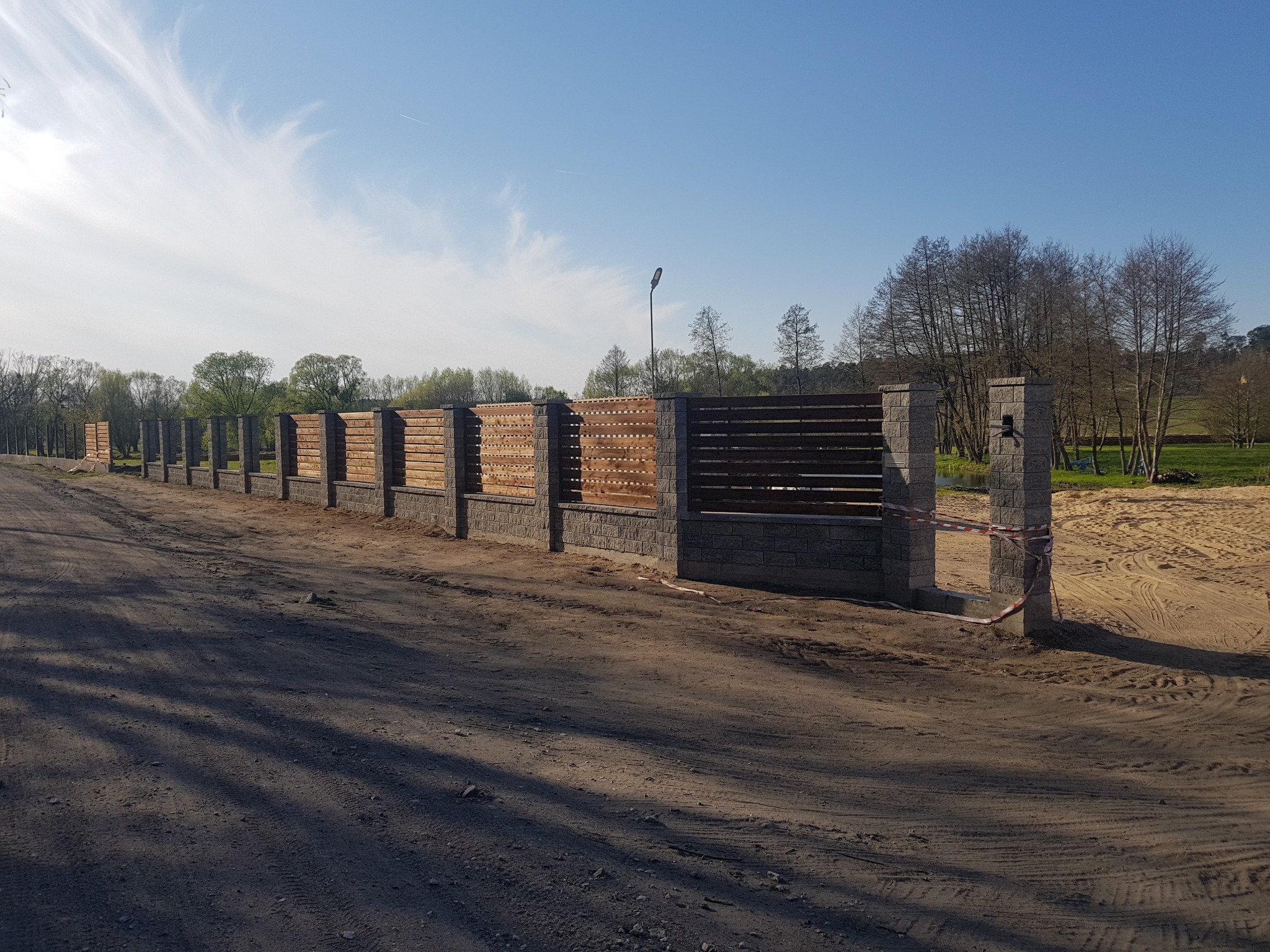 Nowoczesne, poziome ogrodzenie z drewnianymi panelami i kamiennymi słupkami, oddzielające teren budowy od zielonego krajobrazu z drzewami i błękitnym niebem.