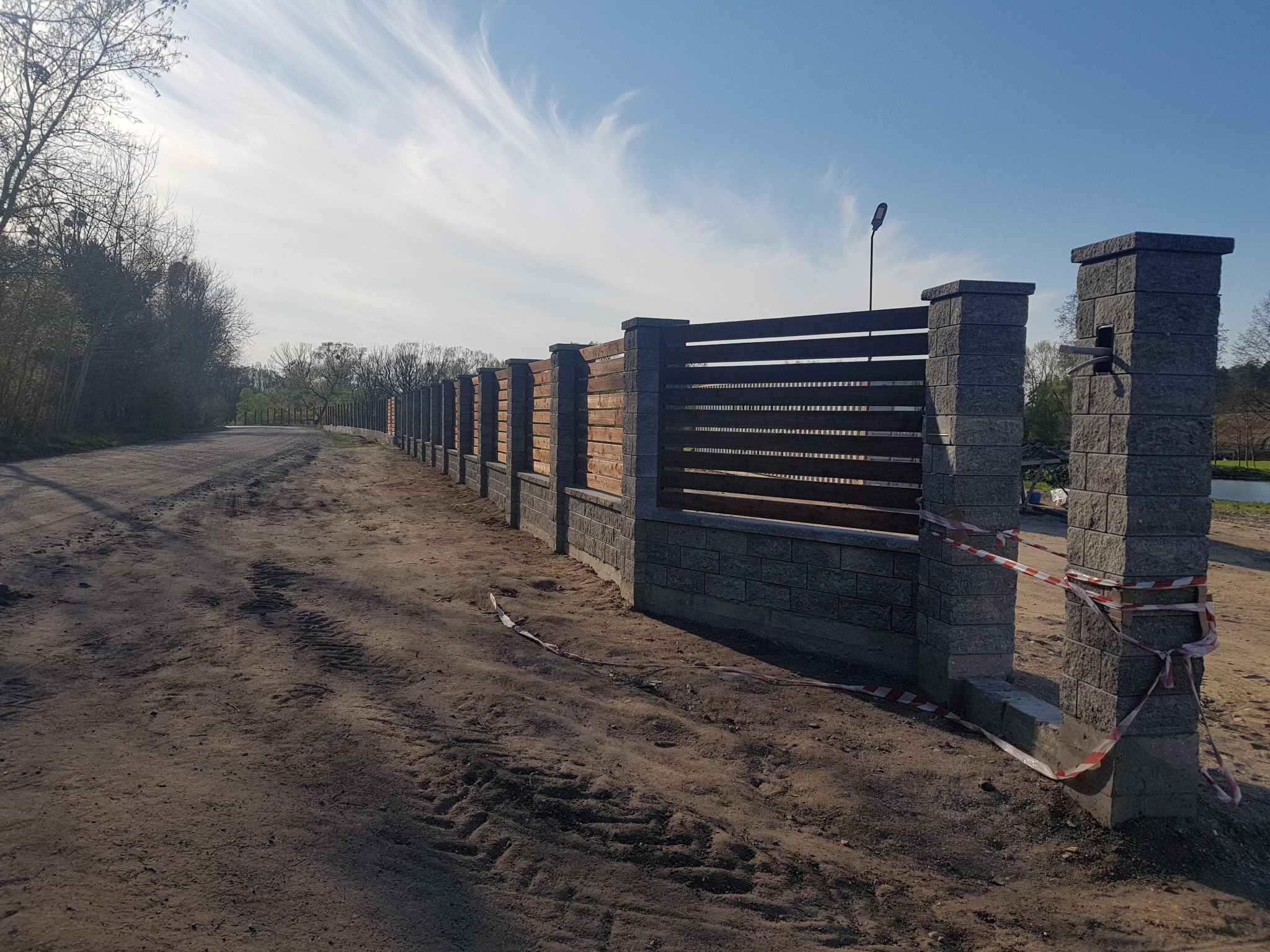 Nowoczesne ogrodzenie posesji z szarych, betonowych słupków i drewnianych paneli, widok na długi odcinek ogrodzenia wzdłuż polnej drogi, z taśmą ostrzegawczą przy wejściu.