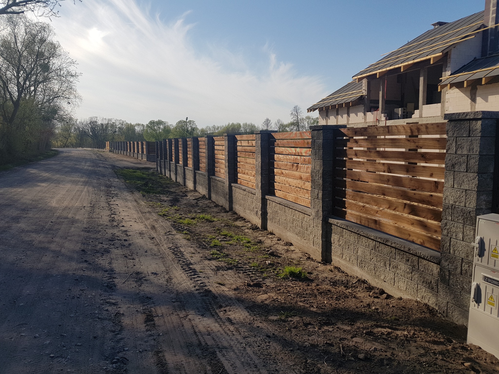 Długie ogrodzenie z drewnianymi panelami i kamiennymi słupkami, biegnące wzdłuż polnej drogi, z widocznym w tle budynkiem w trakcie budowy.