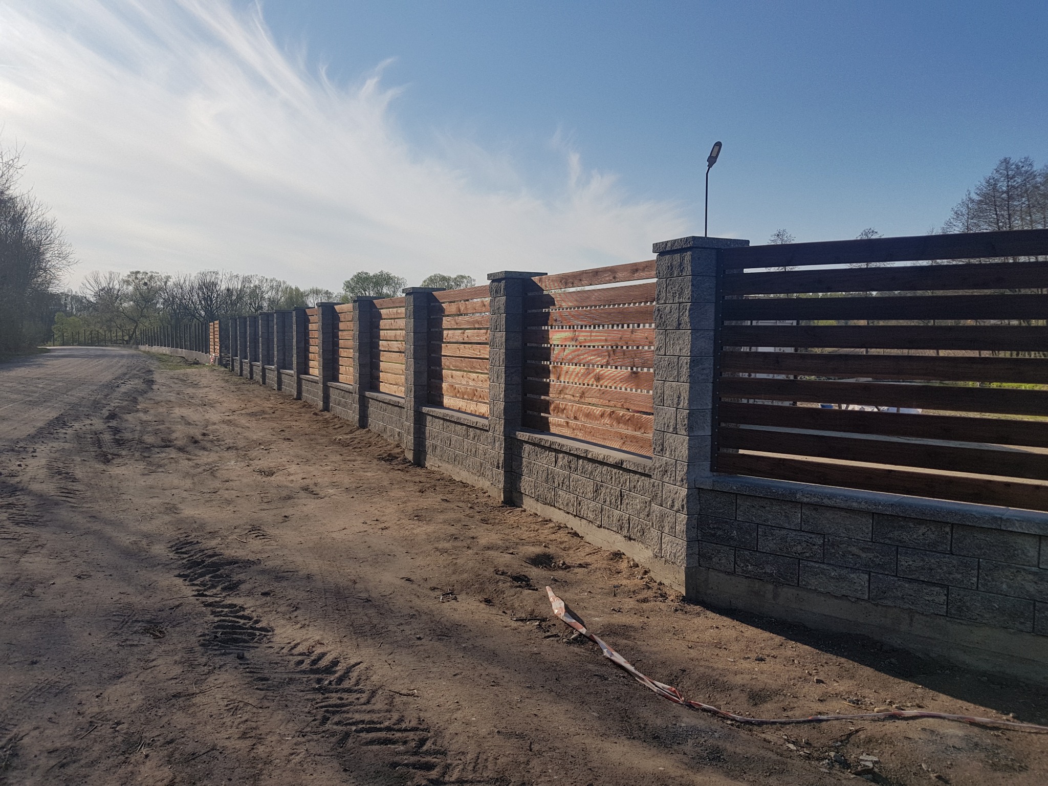 Długi, nowoczesny płot z szarych bloczków betonowych i poziomych, brązowych desek, oświetlony latarnią, biegnący wzdłuż polnej drogi pod błękitnym niebem z delikatnymi chmurami.