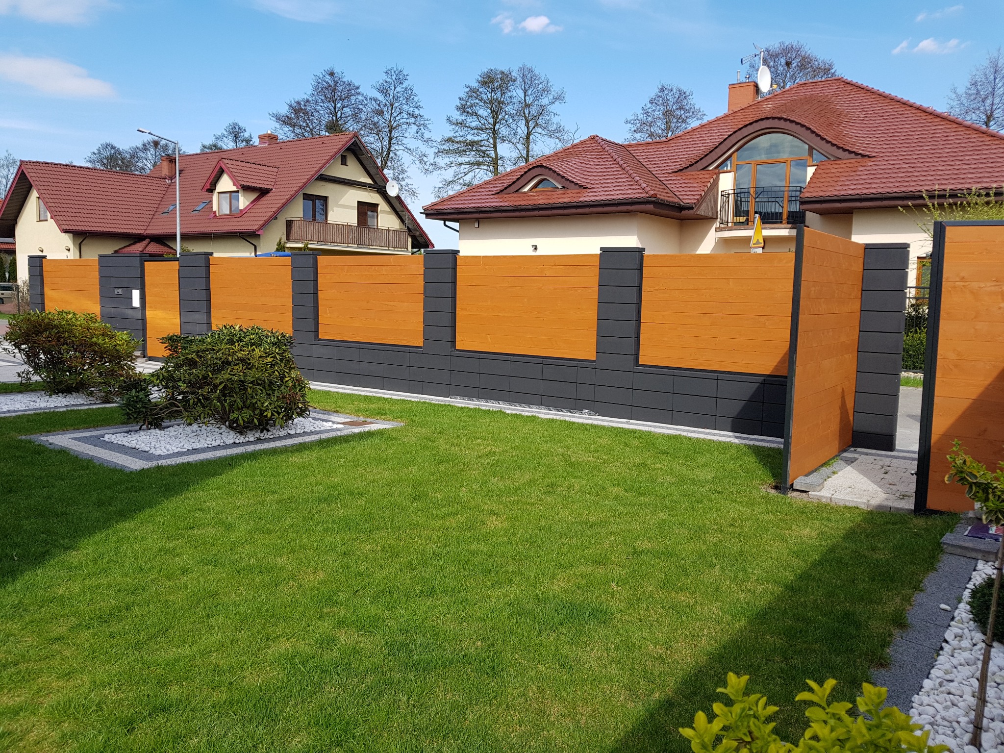 Nowoczesne ogrodzenie panelowe z drewnianymi przęsłami i betonowymi słupkami w kolorze grafitowym, na tle zadbanego trawnika i dwóch domów z czerwoną dachówką.