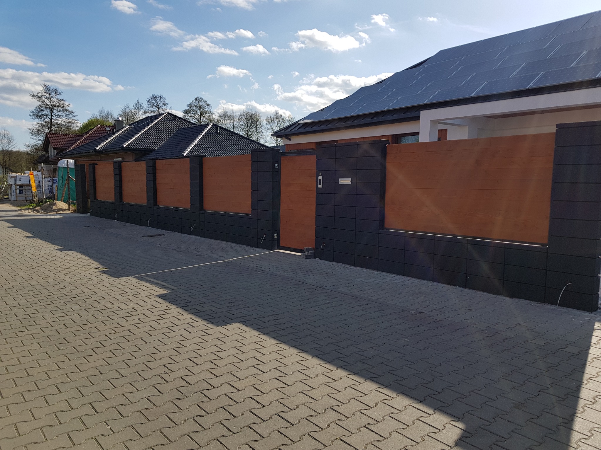 Nowoczesne ogrodzenie panelowe z elementami imitującymi drewno, połączone z ciemnoszarą kostką brukową i domem z panelami słonecznymi na dachu w tle.