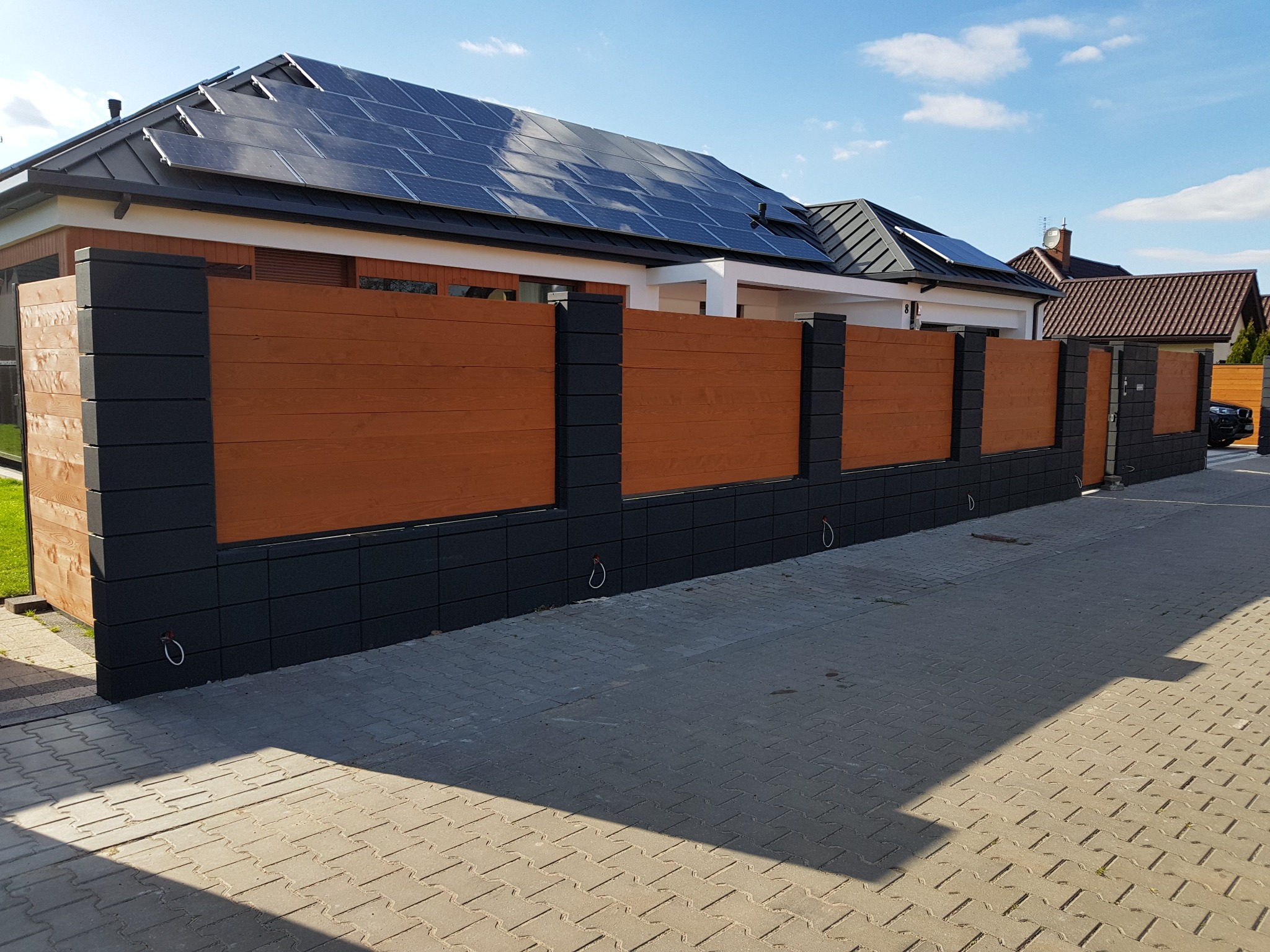 Nowoczesne ogrodzenie panelowe z poziomych desek w kolorze brązowym i słupków z szarego betonu, z widocznymi metalowymi uchwytami, na tle domu z panelami słonecznymi na dachu i brukowanym podjazdem.