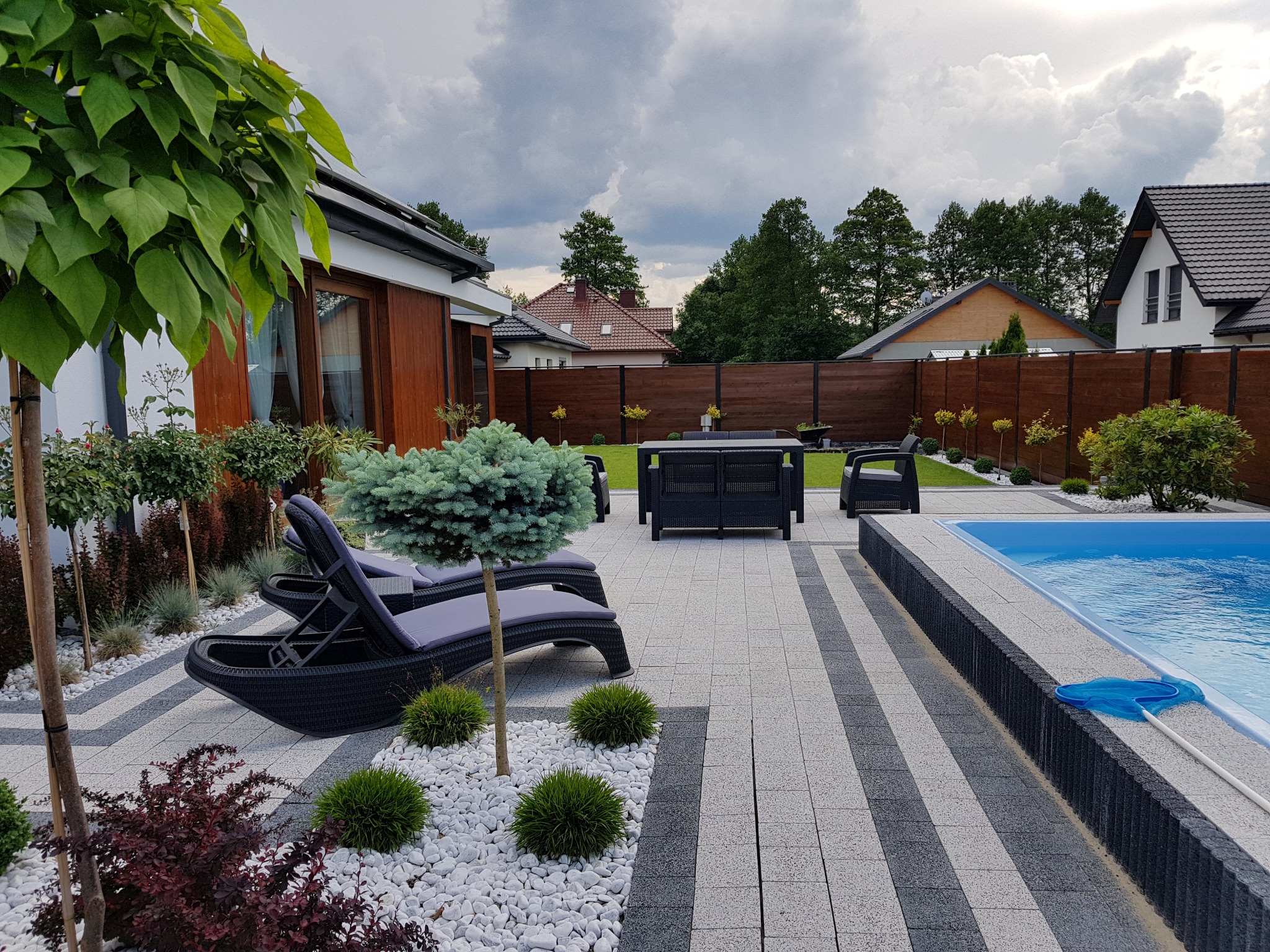 Nowoczesny ogród z basenem w Głogowie: szare i białe płytki chodnikowe, leżaki z poduszkami, zadbany trawnik i drewniane ogrodzenie w tle.