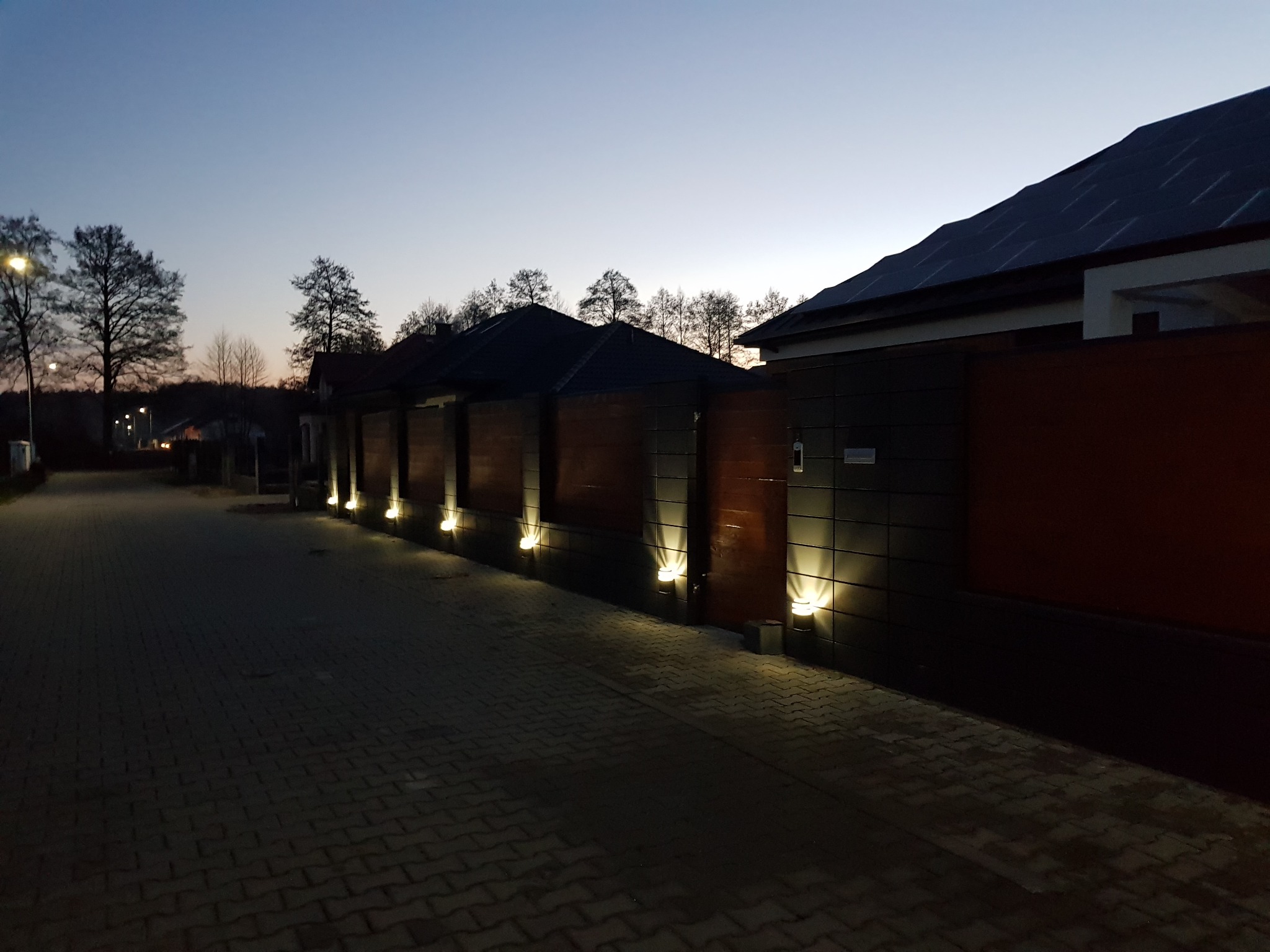 Nowoczesne ogrodzenie posesji z elementami drewna i subtelnym oświetleniem punktowym wzdłuż chodnika z kostki brukowej, sfotografowane o zmierzchu.