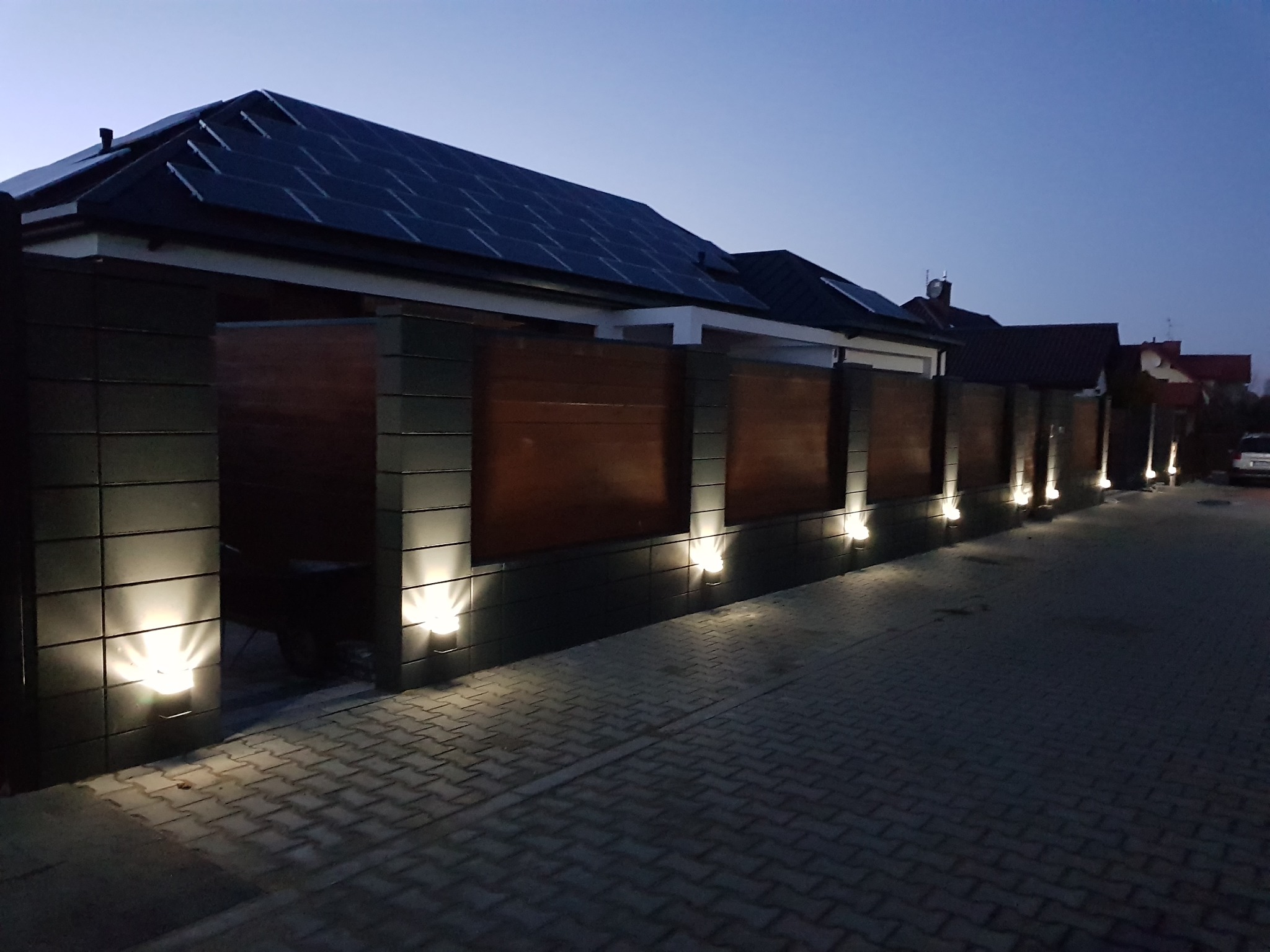 Nowoczesne ogrodzenie posesji z drewnianymi panelami i podświetleniem punktowym, widok wieczorny na tle domu z panelami słonecznymi na dachu.