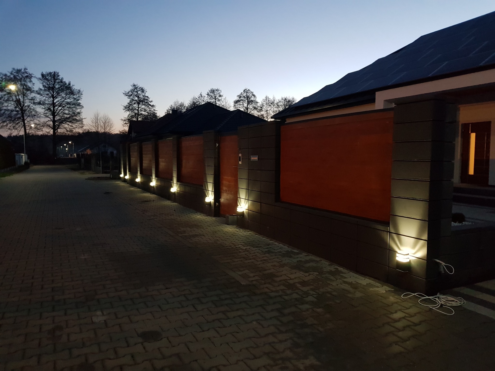 Nowoczesne ogrodzenie posesji z paneli drewnianych i słupków z szarej kostki, oświetlone punktowymi lampami wbudowanymi w słupki, widok na posesję o zmierzchu.