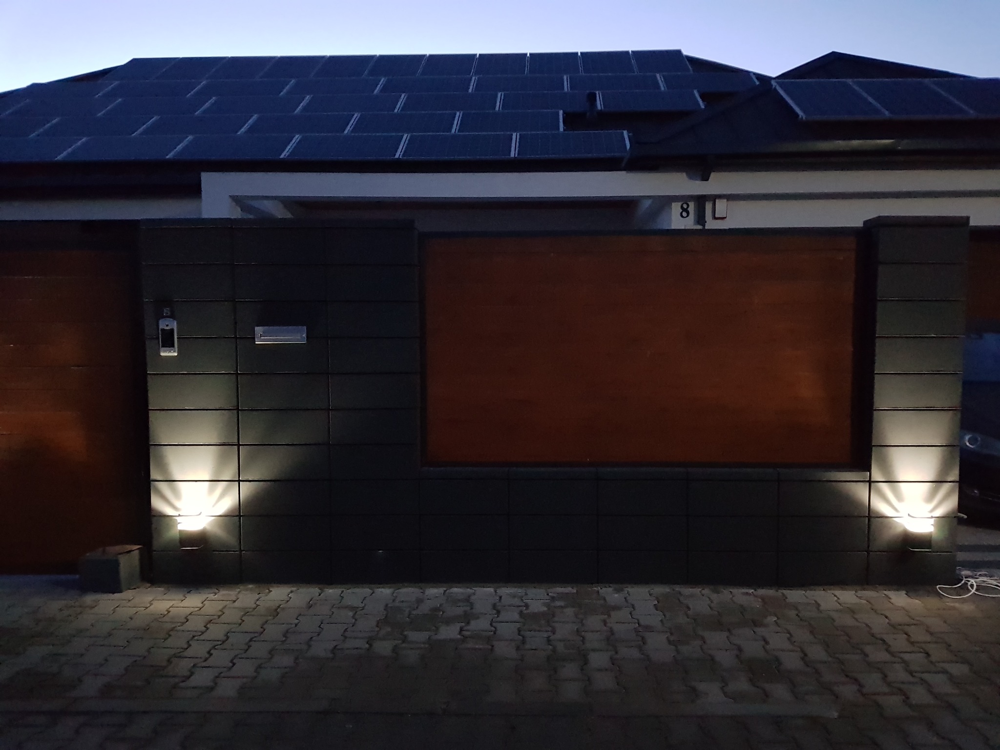 Nowoczesne ogrodzenie panelowe z elementami drewnianymi i oświetleniem punktowym, na tle domu z panelami słonecznymi na dachu i kostką brukową.