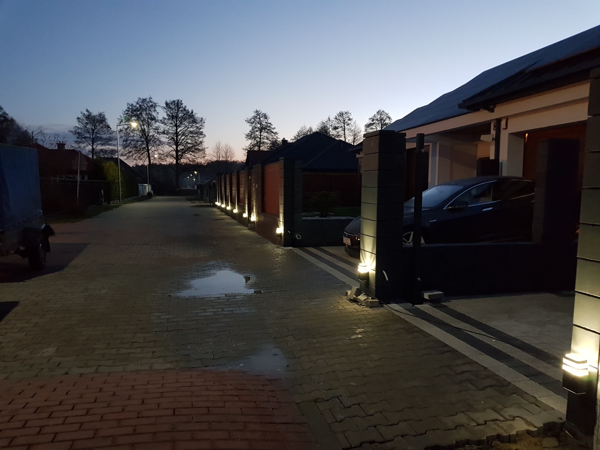 Nowoczesne ogrodzenie z ceglanymi przęsłami i oświetleniem punktowym wzdłuż posesji, z widocznym fragmentem elewacji domu i zaparkowanym samochodem w tle, sfotografowane o zmierzchu.