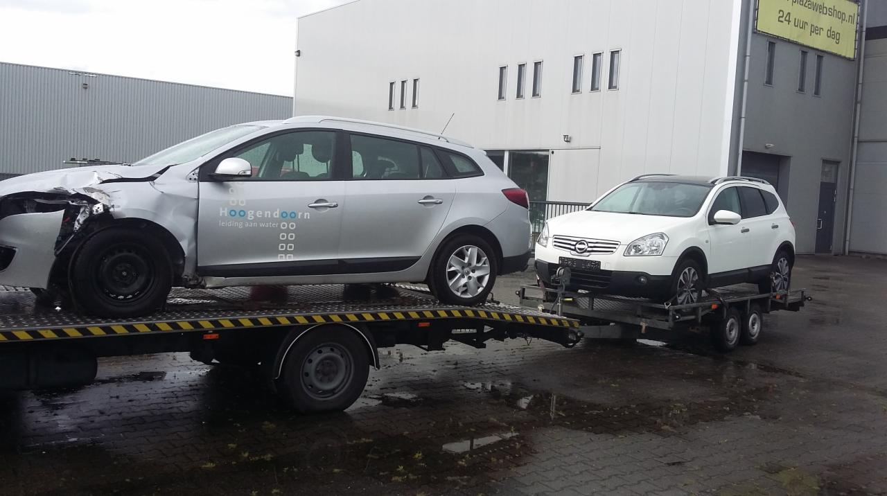 Srebrny samochód z uszkodzonym przodem załadowany na lawetę, za nim biały samochód na przyczepie, budynek w tle.