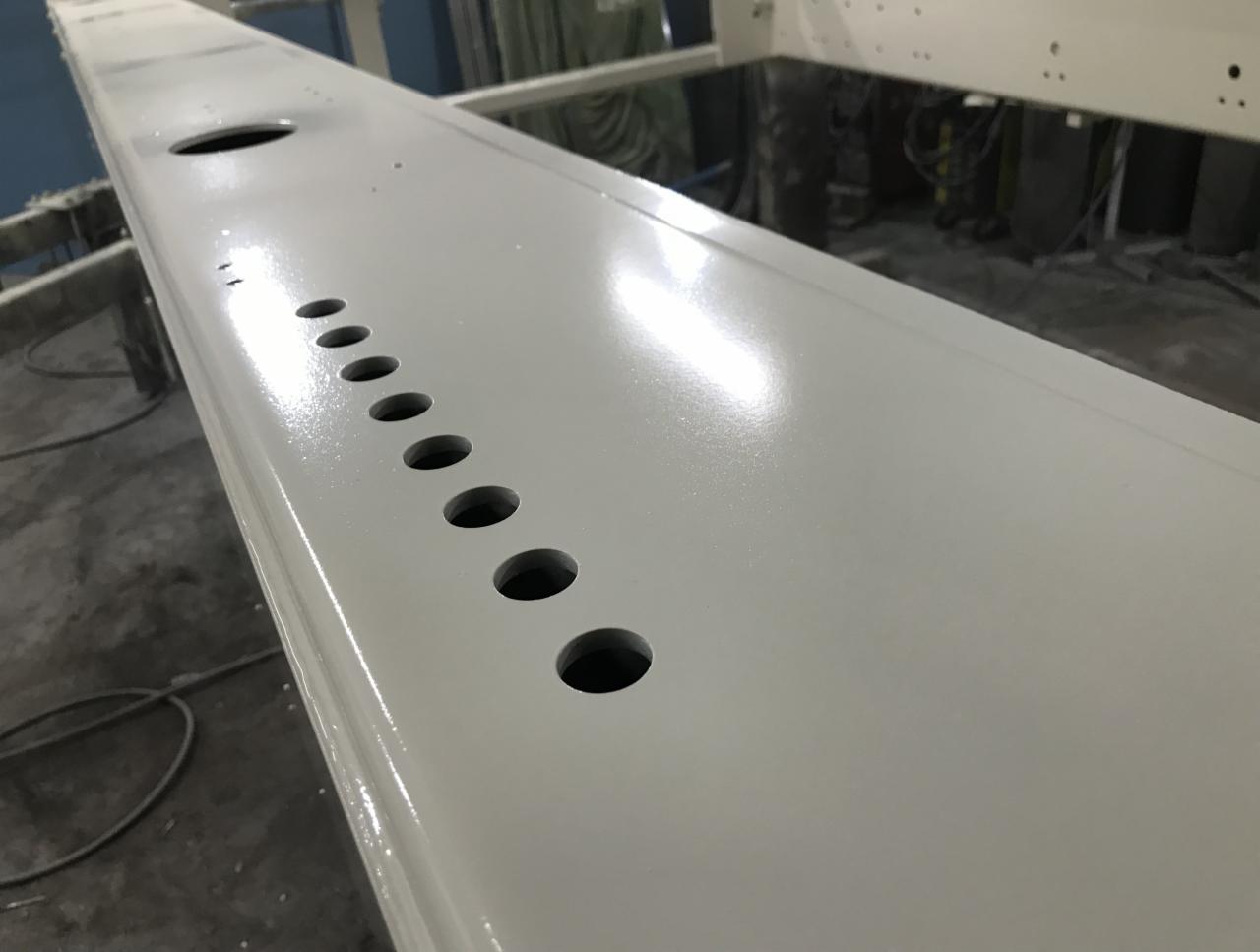 Szlifowany, pomalowany na jasny kolor metalowy profil z rzędem okrągłych otworów, widoczny w perspektywie, z elementami konstrukcji w tle.