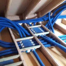 Niebieskie, karbowane przewody wentylacyjne prowadzone w drewnianej konstrukcji stropu poddasza, widoczne elementy mocujące i izolacja.