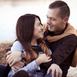 Uśmiechnięta para zakochanych siedzi blisko siebie na tle jesiennego krajobrazu; mężczyzna obejmuje kobietę, oboje patrzą na siebie z czułością. Na pierwszym planie widoczny damski zegarek na ręce.