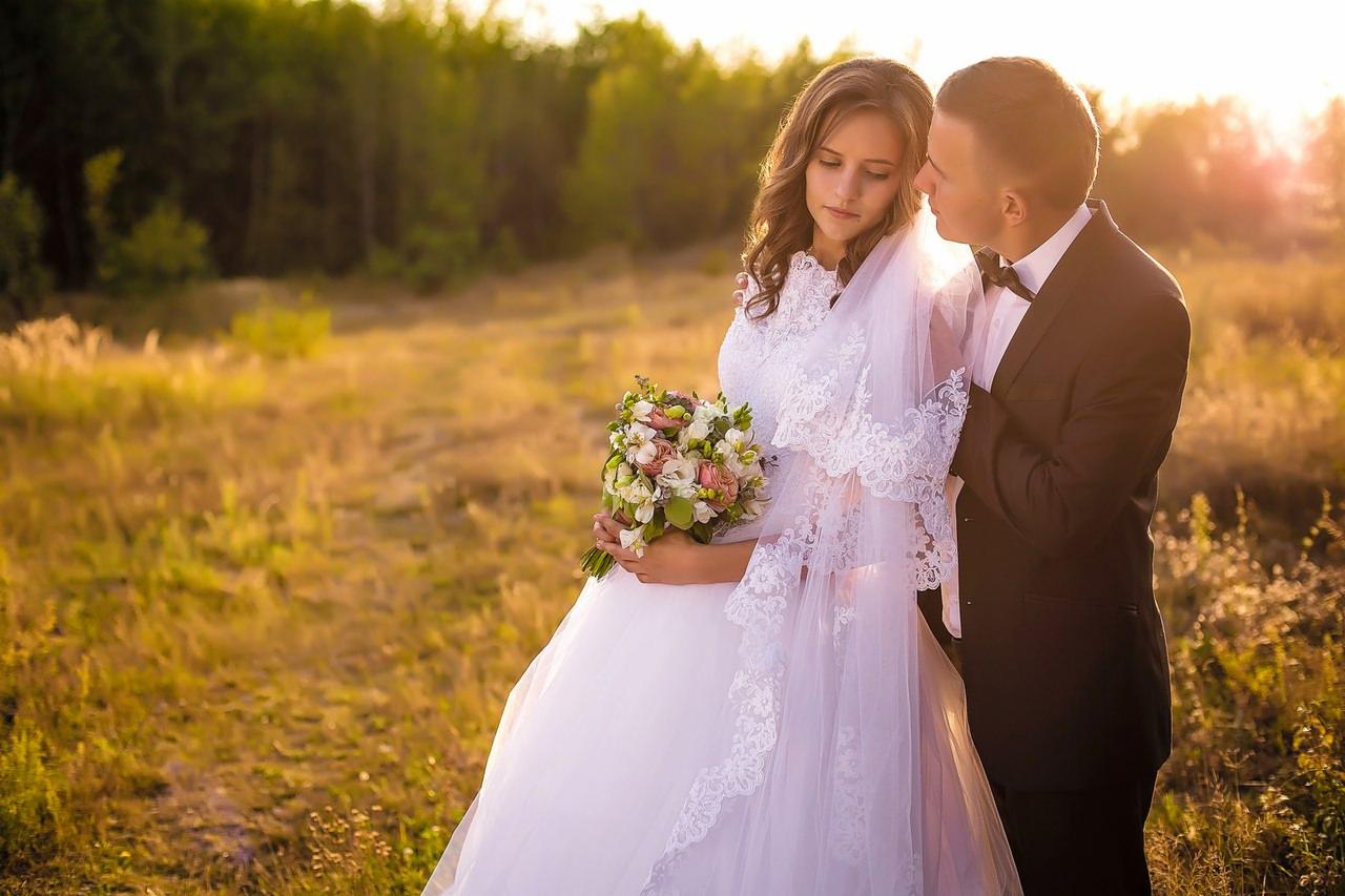 Para młoda w plenerze: panna młoda w białej sukni z koronkowym welonem trzyma bukiet, pan młody w czarnym smokingu delikatnie obejmuje ją, ciepłe światło zachodzącego słońca w tle.