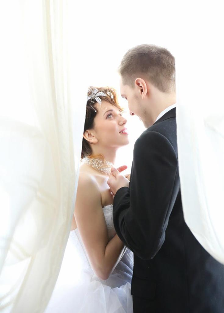 Panna młoda w tiulowej sukni i zdobionej opasce, spogląda z uśmiechem na pana młodego w czarnym garniturze, oboje stoją za delikatną, białą zasłoną.