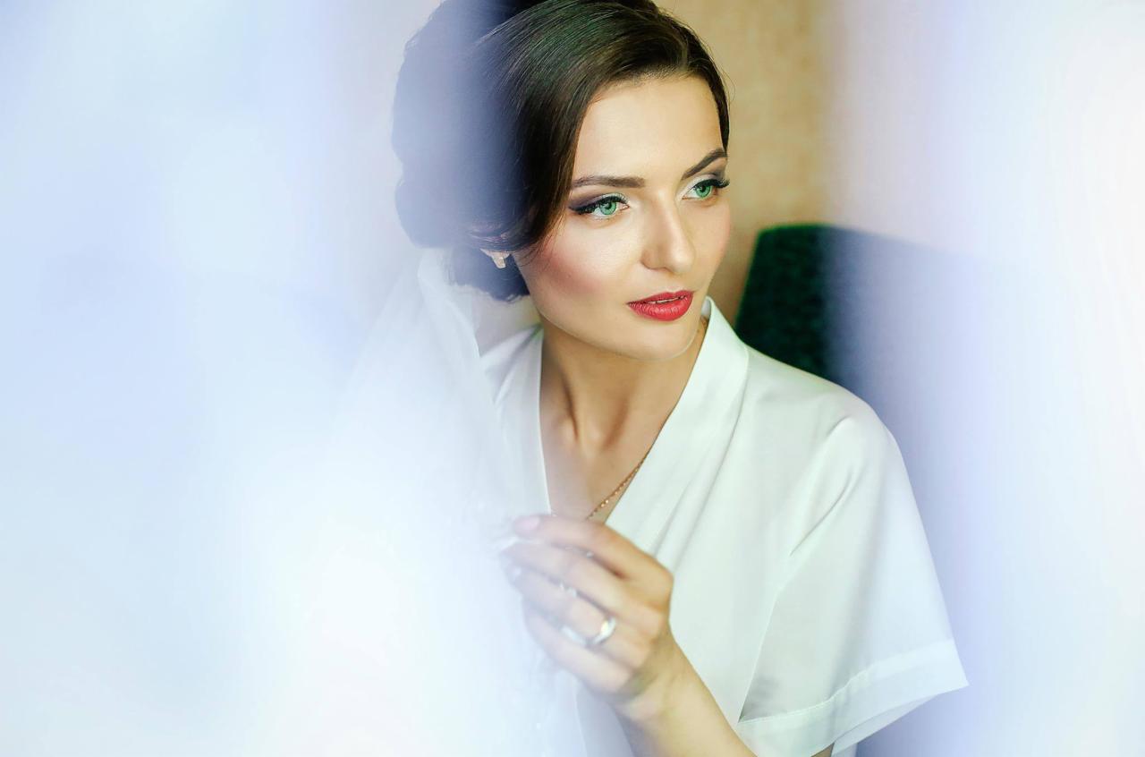 Panna młoda w białym szlafroku, poprawia welon, makijaż z zielonymi oczami i czerwoną szminką, delikatna biżuteria.
