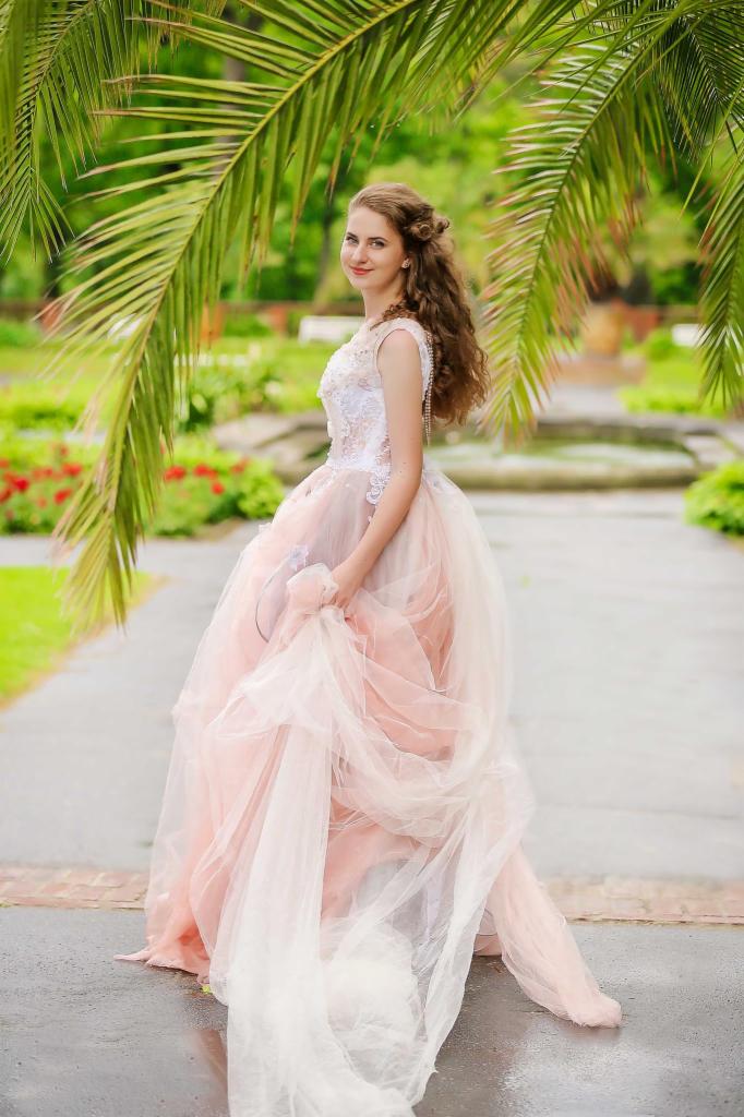 Uśmiechnięta kobieta w długiej, balowej sukni w odcieniach różu i bieli pozuje w ogrodzie z palmami i fontanną w tle.