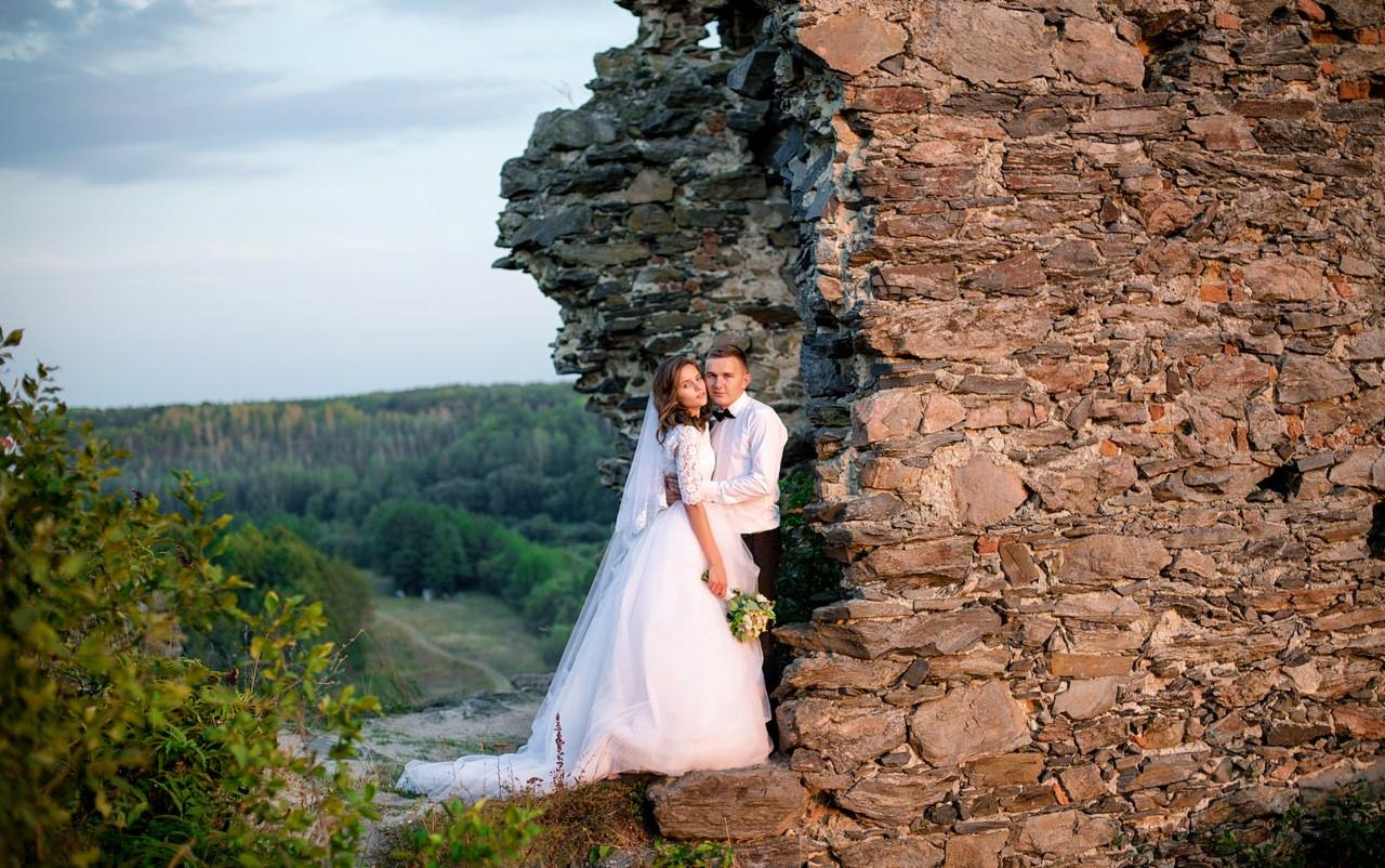 Para młoda w objęciach na tle kamiennych ruin zamku i zielonego lasu, ona w białej sukni z koronkowymi rękawami i welonem, on w białej koszuli i czarnej muszce.