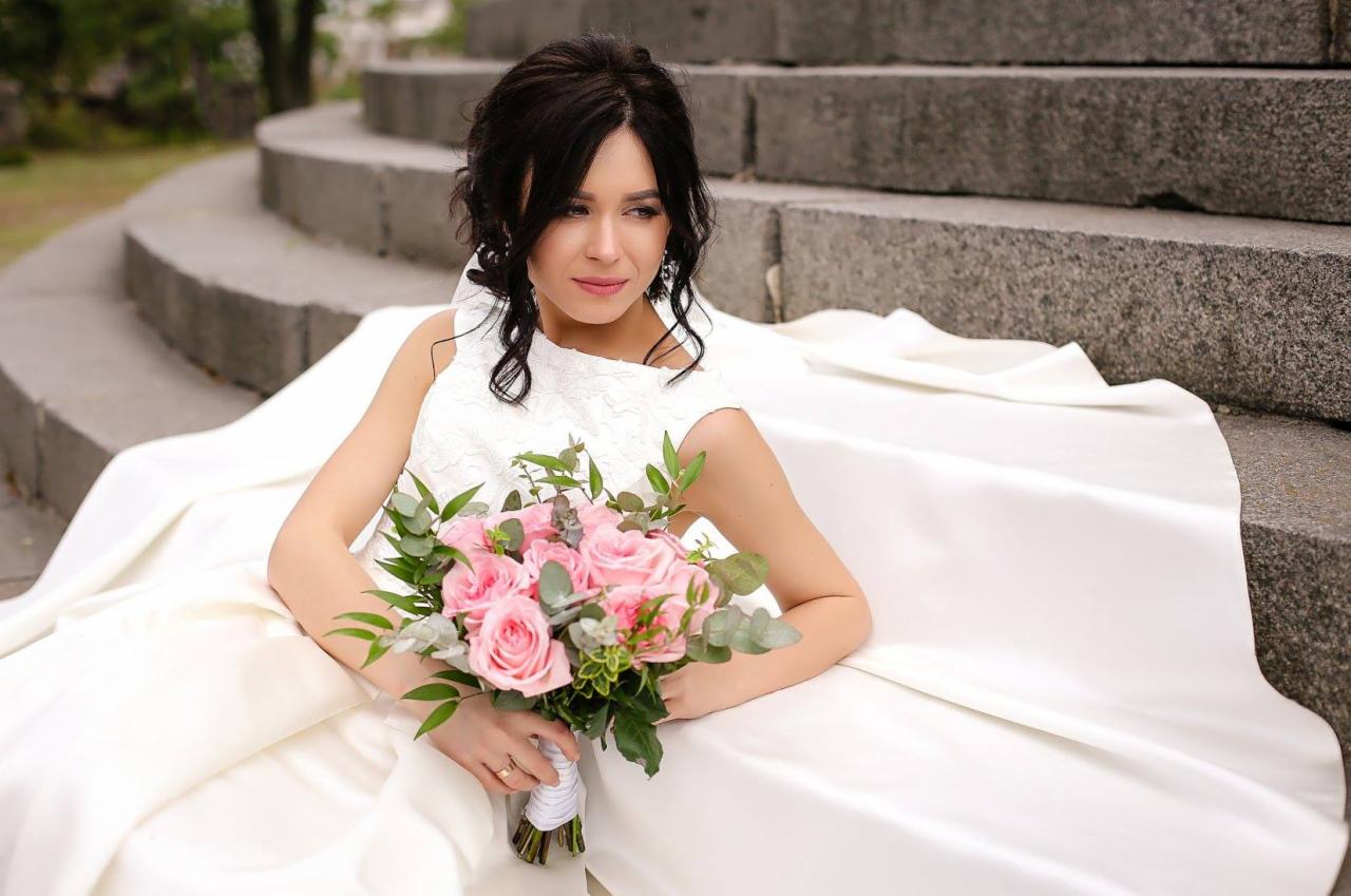 Panna młoda w białej sukni siedzi na schodach, trzyma bukiet różowych róż i spogląda w bok.