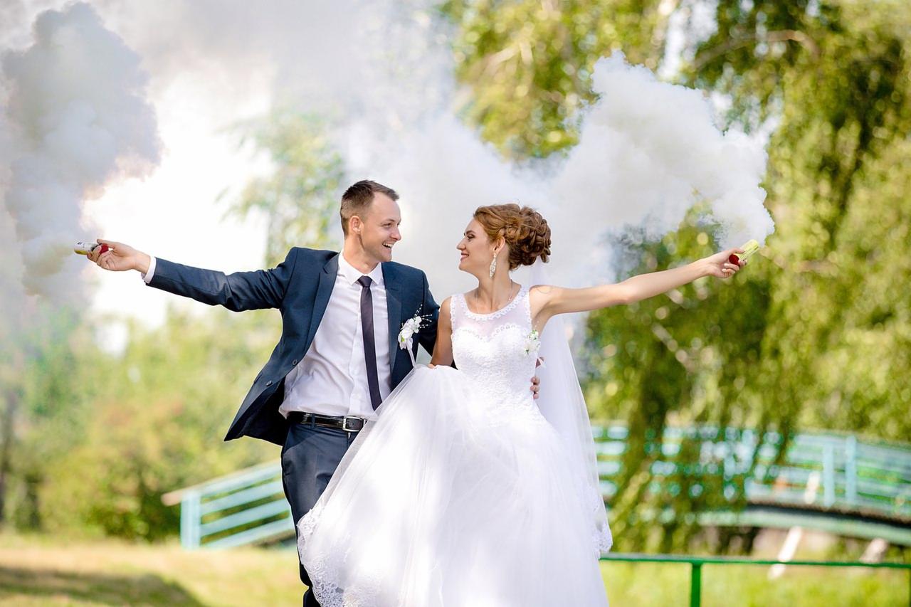 Uśmiechnięta para młoda, trzymająca w dłoniach odpalone race dymne na tle zieleni.