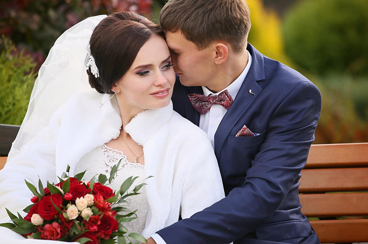 Panna młoda w białym futerku i welonie, trzyma bukiet czerwonych róż, przytulona do pana młodego w granatowym garniturze z bordową muchą, siedzą na drewnianej ławce.