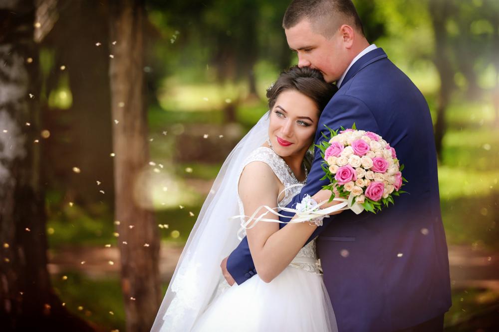 Panna młoda w białej sukni, z bukietem różowych i kremowych róż, obejmowana przez pana młodego w granatowym garniturze na tle rozmytej zieleni drzew i delikatnych świetlnych refleksów.