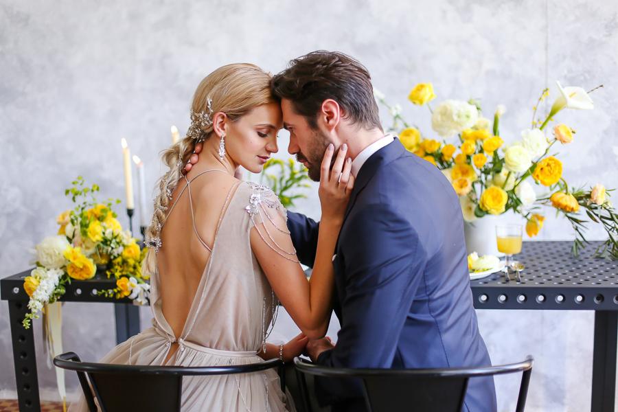 Elegancka para w objęciach przy stole z żółtymi kwiatami i świecami, panna młoda w sukni z odkrytymi plecami i ozdobnymi łańcuszkami.