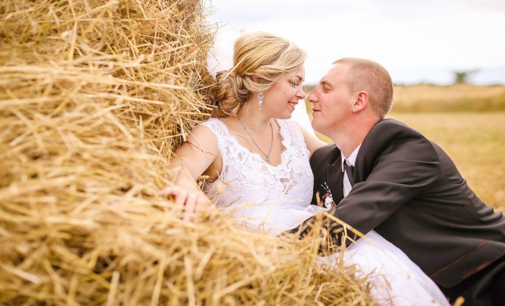Para młoda w strojach ślubnych, siedząca w stogu siana na polu, twarzami blisko siebie, delikatny uśmiech.