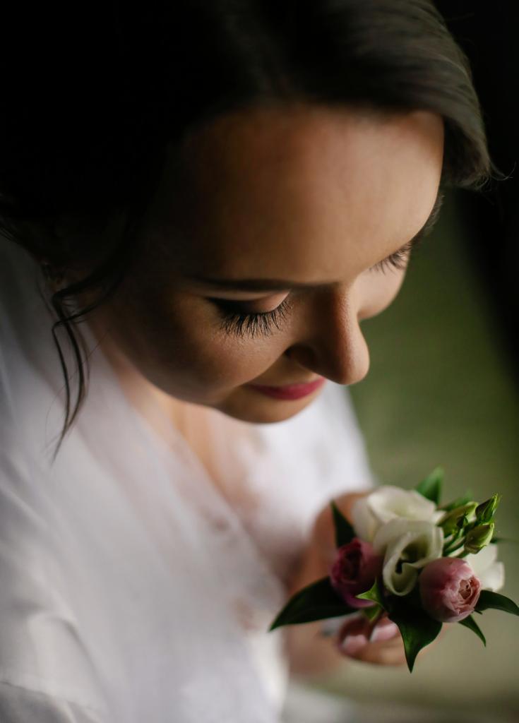 Panna młoda z delikatnym bukiecikiem w dłoniach, spojrzenie skierowane w dół, makijaż z podkreślonymi rzęsami, przygotowania do ślubu.