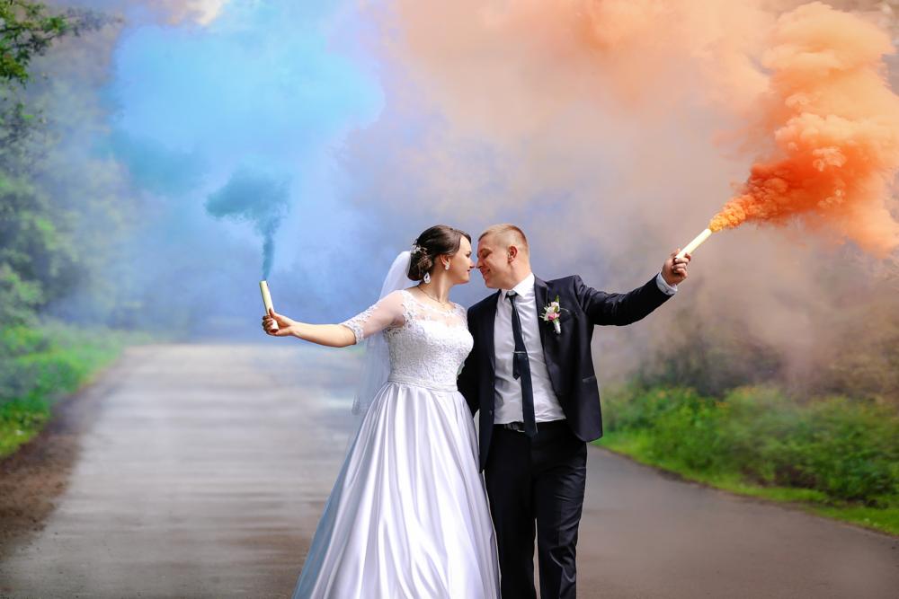 Para młoda na drodze, trzymająca w rękach race dymne w kolorach niebieskim i pomarańczowym, zbliżona twarzami.