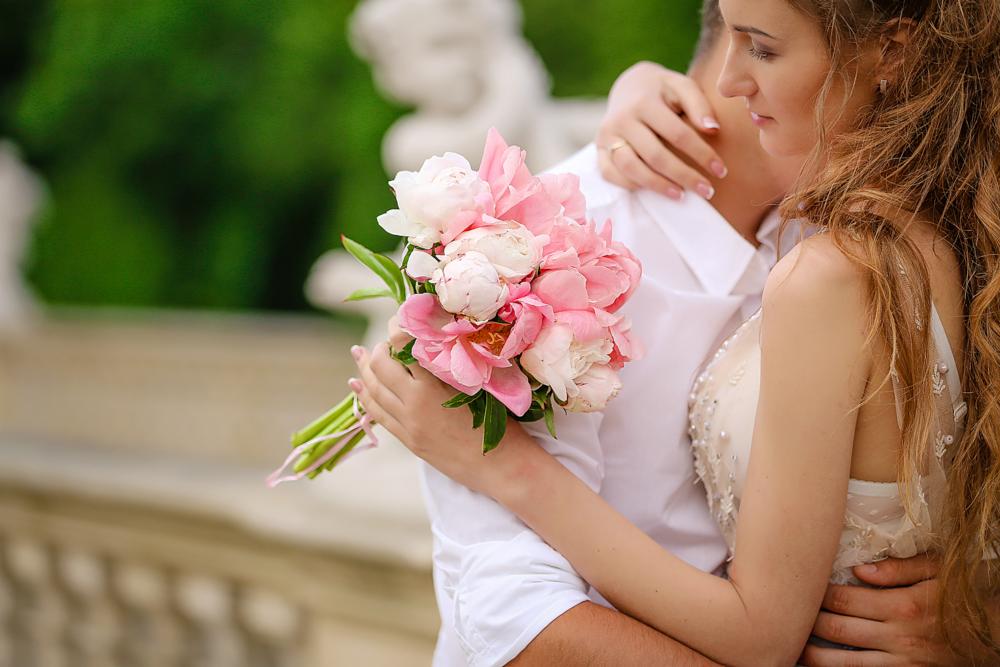 Para młoda obejmująca się w plenerze, panna młoda trzyma bukiet różowych i białych piwonii, w tle rzeźby i zieleń.