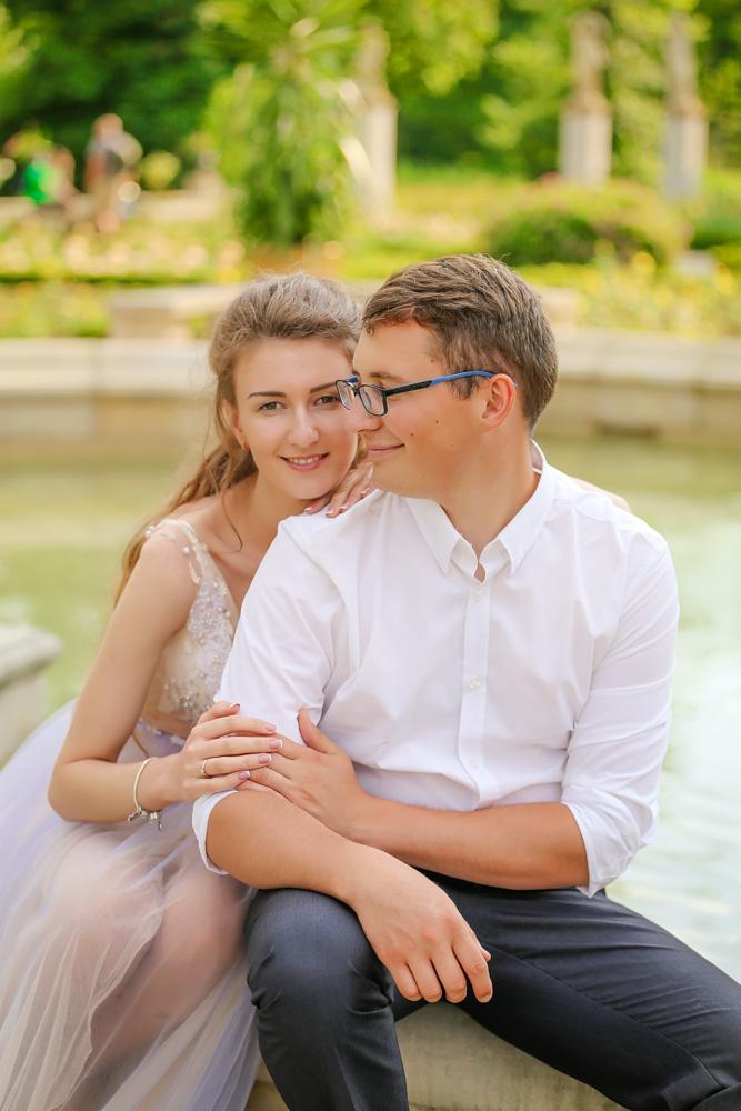 Para w parku podczas sesji narzeczeńskiej: uśmiechnięta kobieta w sukni, opierająca się o mężczyznę w białej koszuli i okularach, na tle fontanny i zieleni.