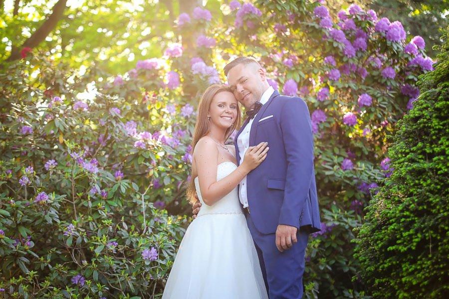 Uśmiechnięta para młoda w objęciach na tle kwitnących rododendronów w słonecznym ogrodzie. Panna młoda w białej sukni bez ramiączek, pan młody w granatowym garniturze i muszce.