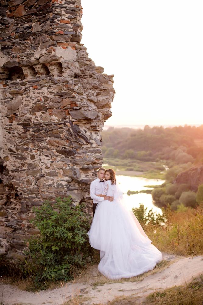 Para młoda w objęciach na tle kamiennych ruin i malowniczego krajobrazu z rzeką w oddali, ujęcie z lekkim zachodem słońca.