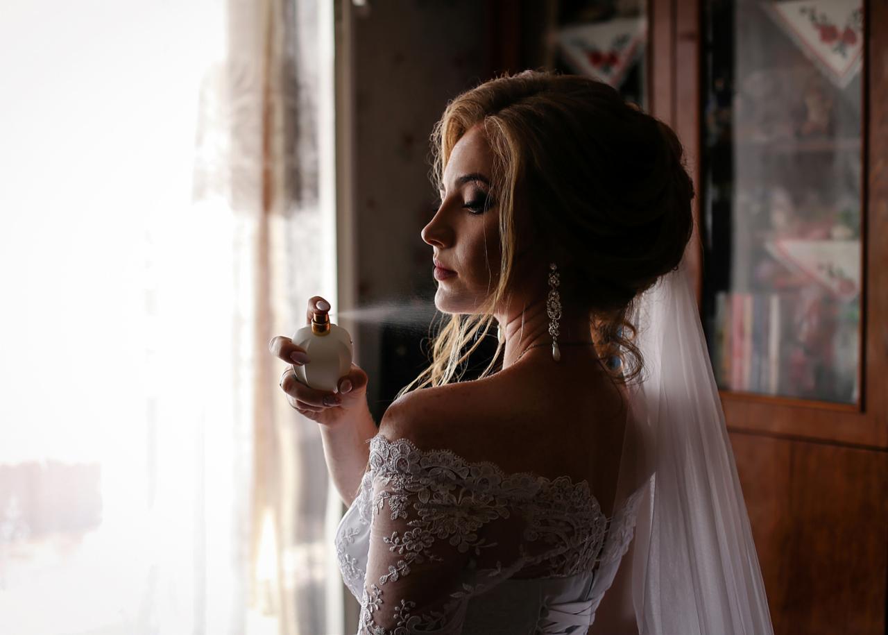 Panna młoda w koronkowej sukni, z welonem, aplikuje perfumy przed lustrem, długie kolczyki z perłą